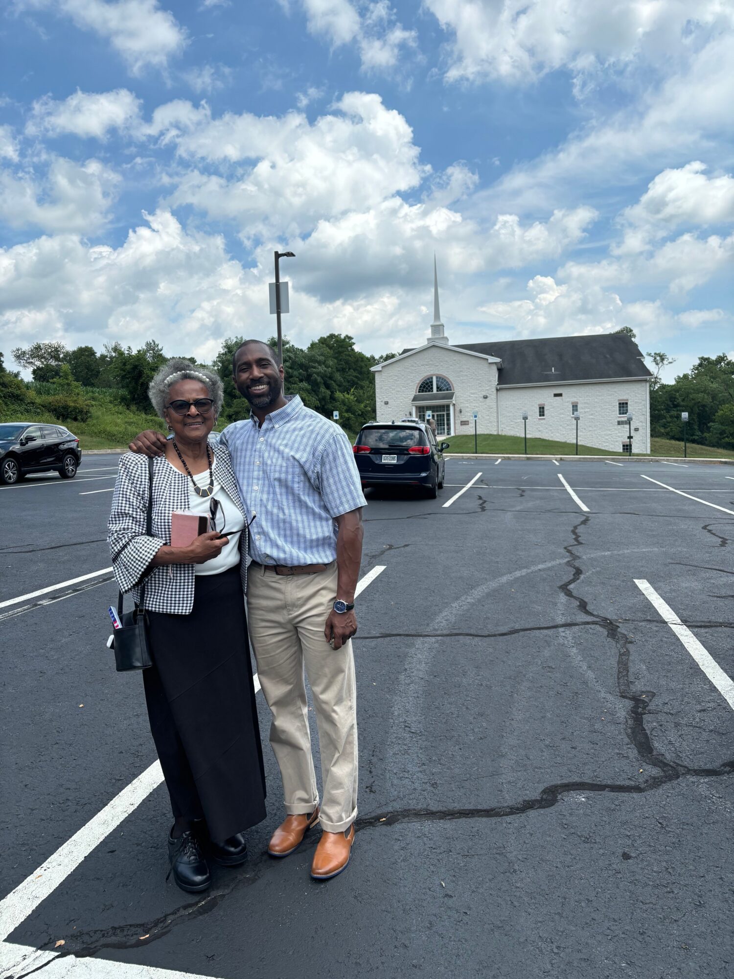 Two people standing in a parking lot with a building and a car in the background, under a partly cloudy sky.