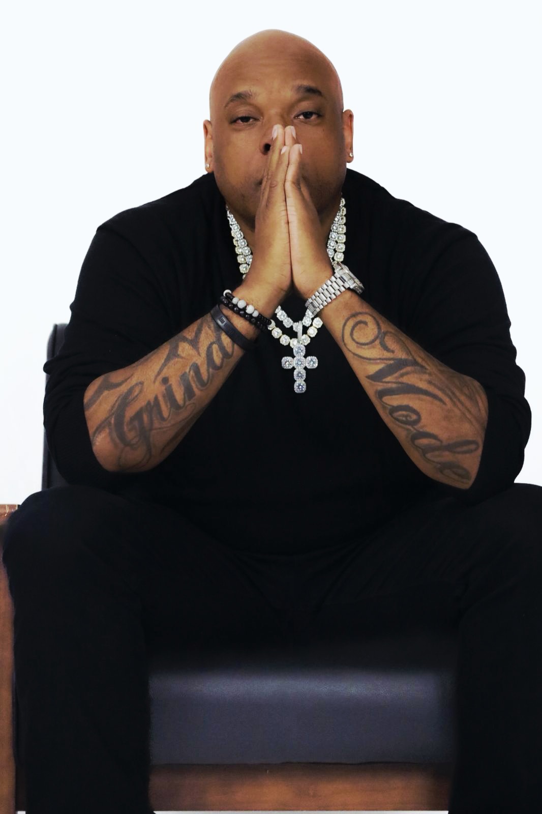 Man with tattoos, wearing a black shirt and jewelry, sitting with hands pressed together in front of face.