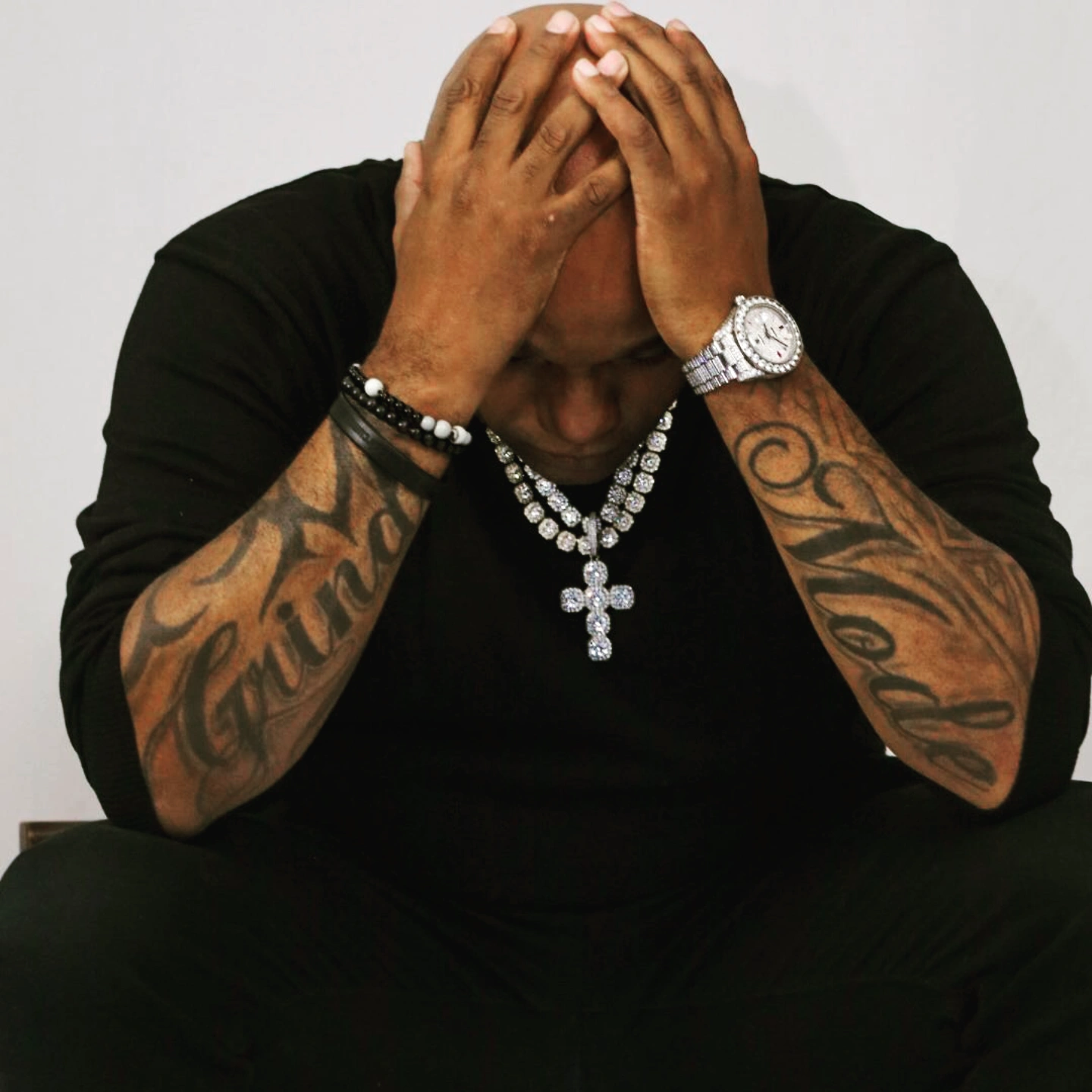 Person with head in hands, wearing black clothing, jewelry, and tattoos on arms, against a plain background.