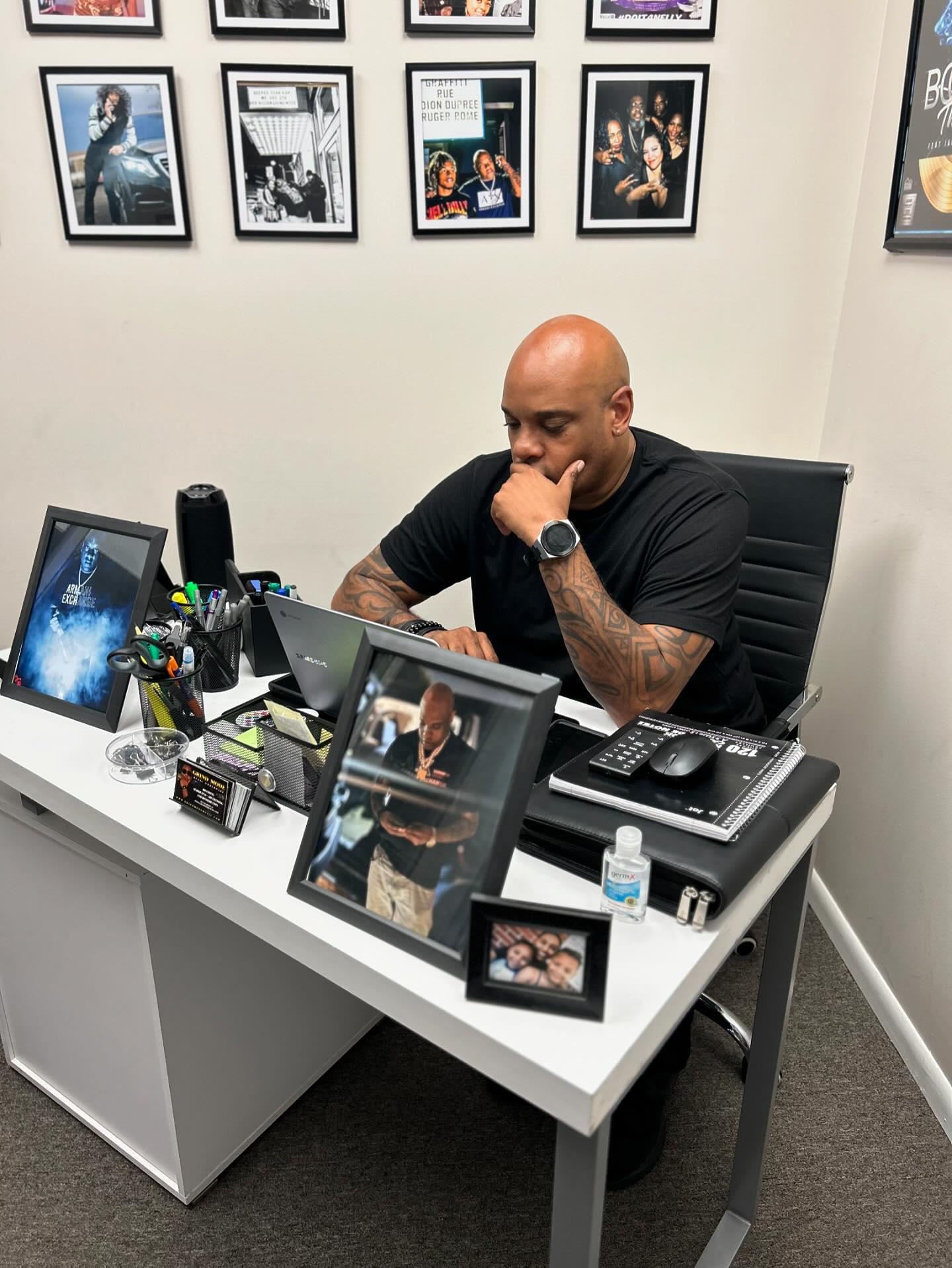 Bald man with tattoos sitting at a desk, working on a laptop, surrounded by framed photos and office supplies.