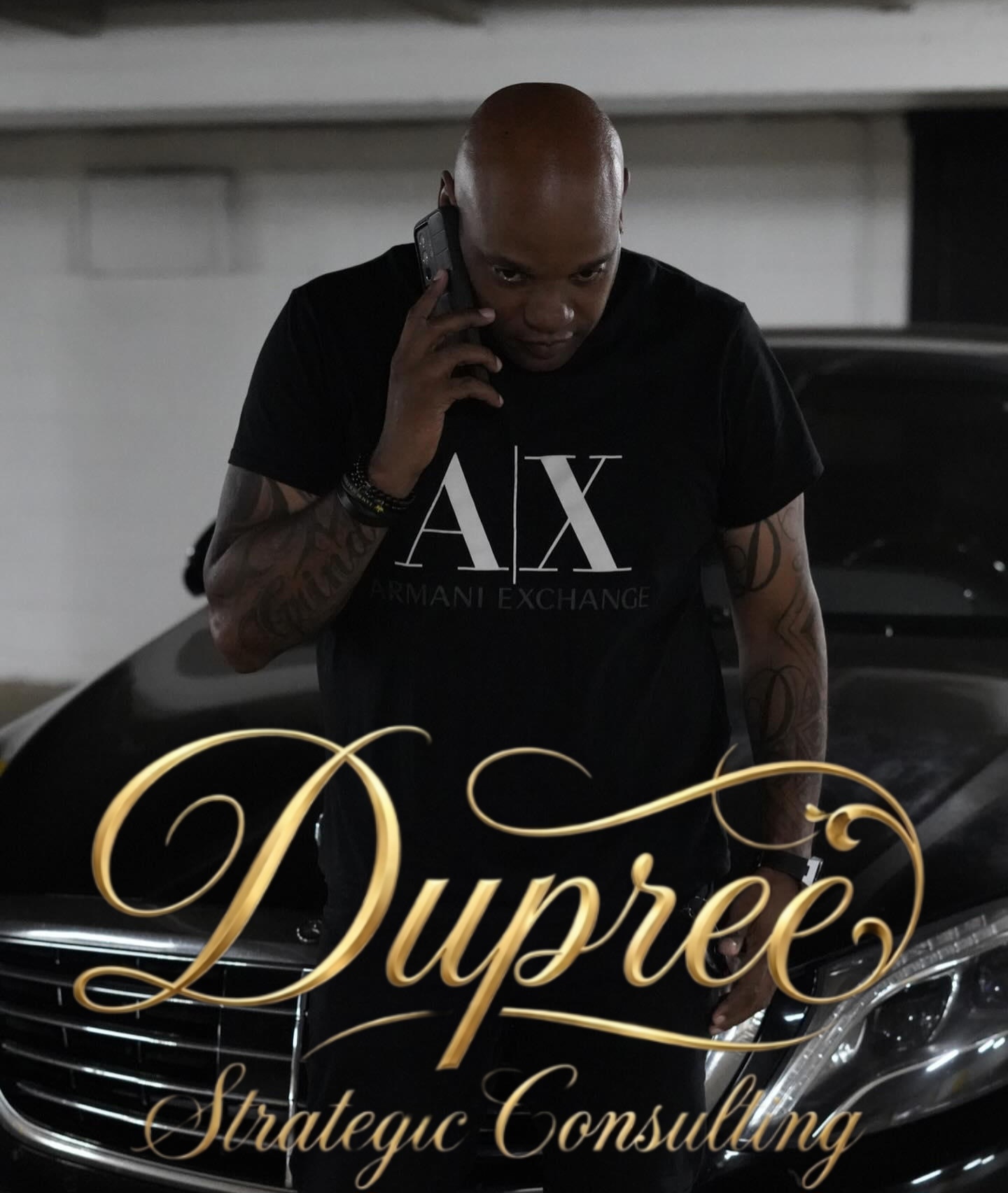 Man with a shaved head talking on a phone, wearing a black Armani Exchange shirt, standing in front of a car.