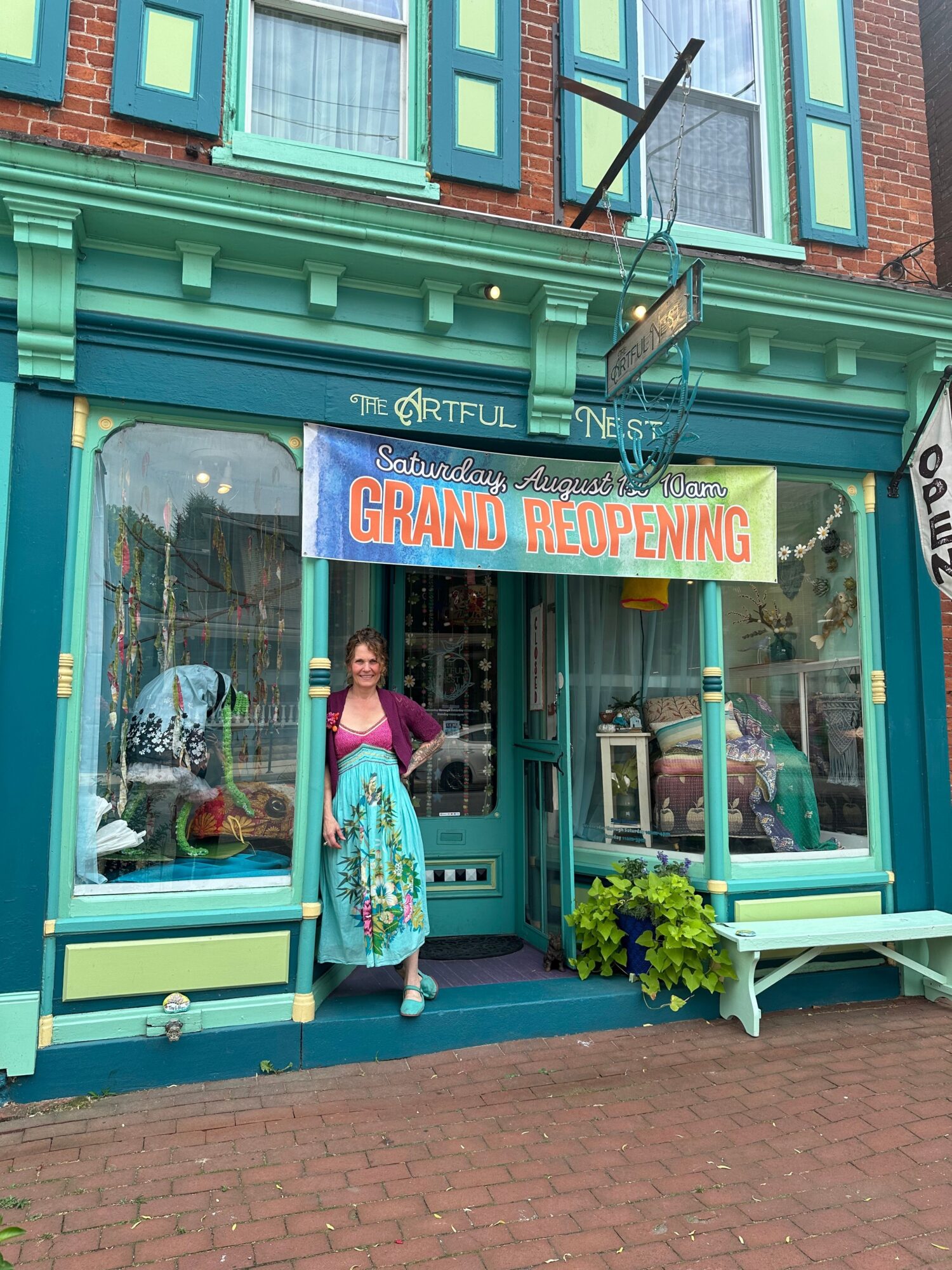 Person standing outside a storefront with a sign that reads 'Grand Reopening,' a potted plant, and a glass door.