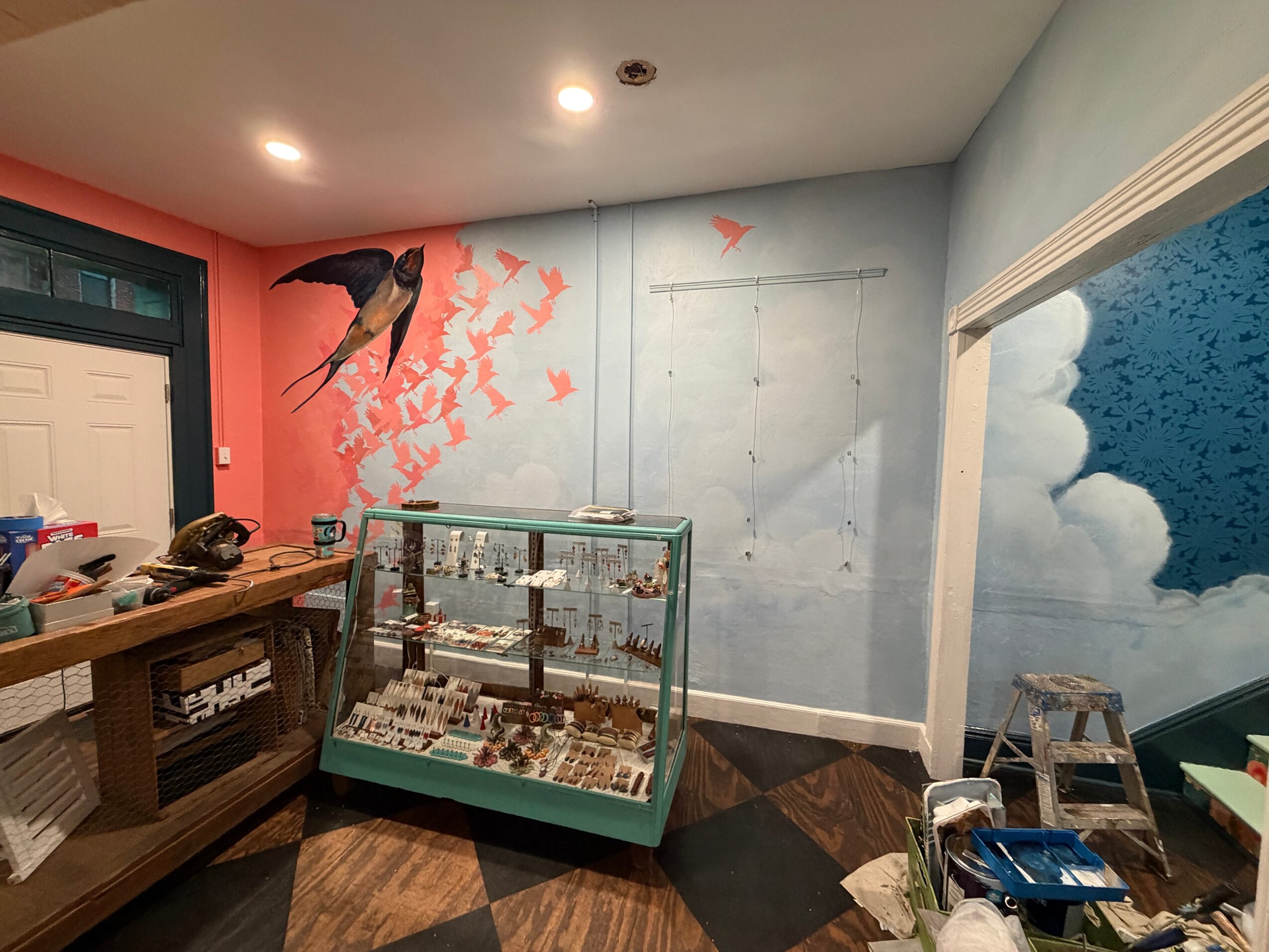 Room with bird mural, display case, and partially painted wall, featuring a ladder and tools, with a door on the left.