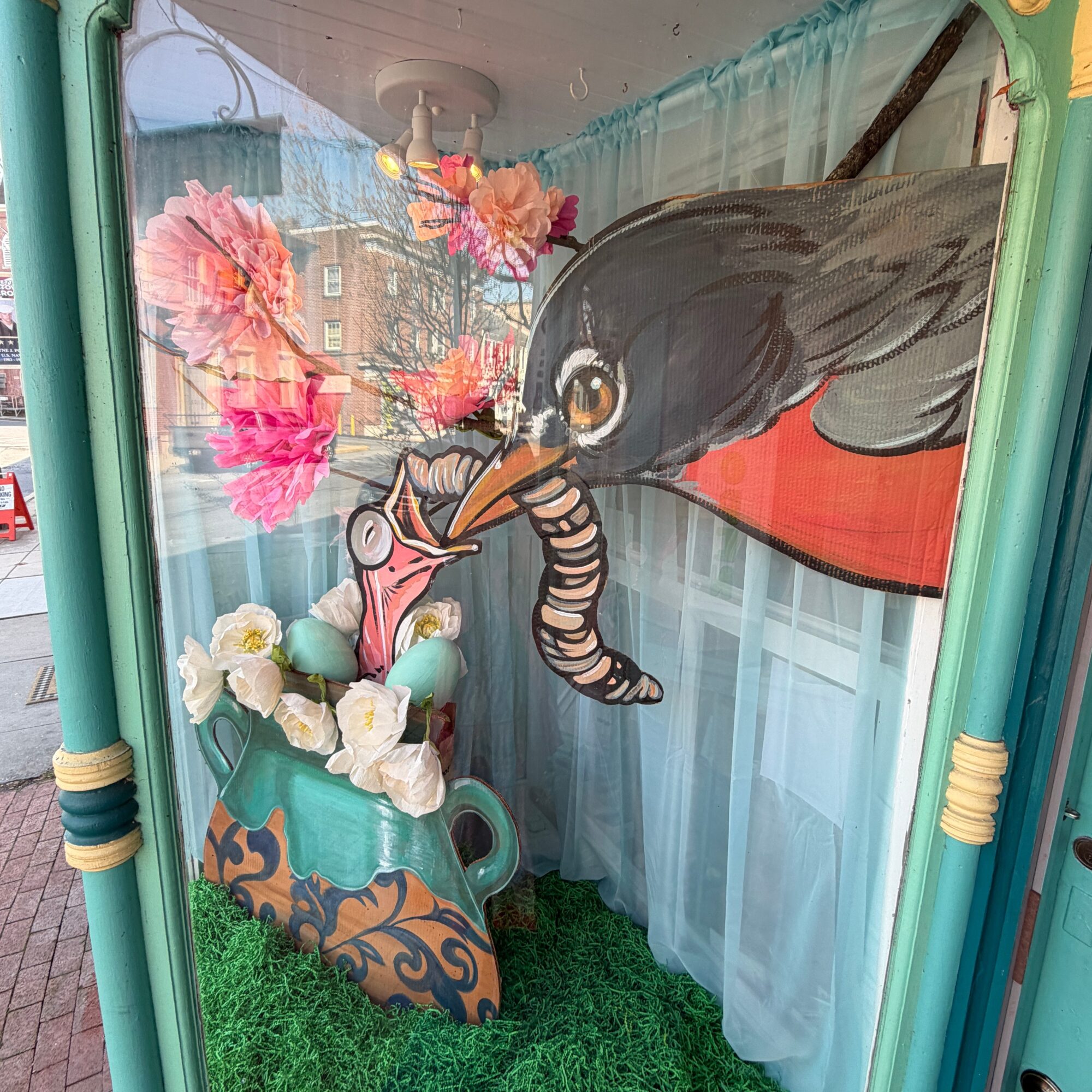 Decorative display with a large bird, flowers, and a teapot, set against a window with curtains and a city view.