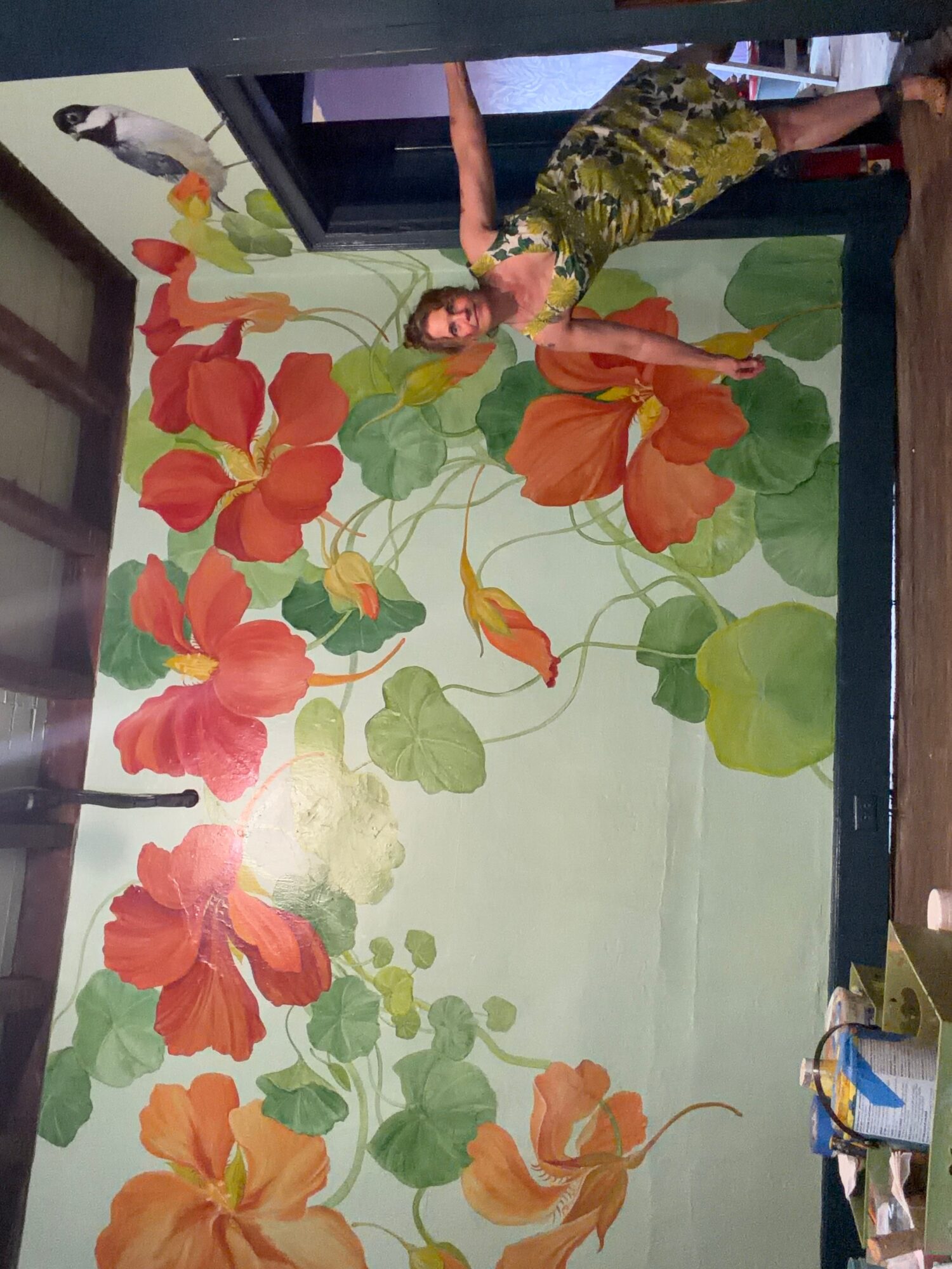Person standing next to a large floral mural with orange and red flowers and green leaves on a wall.