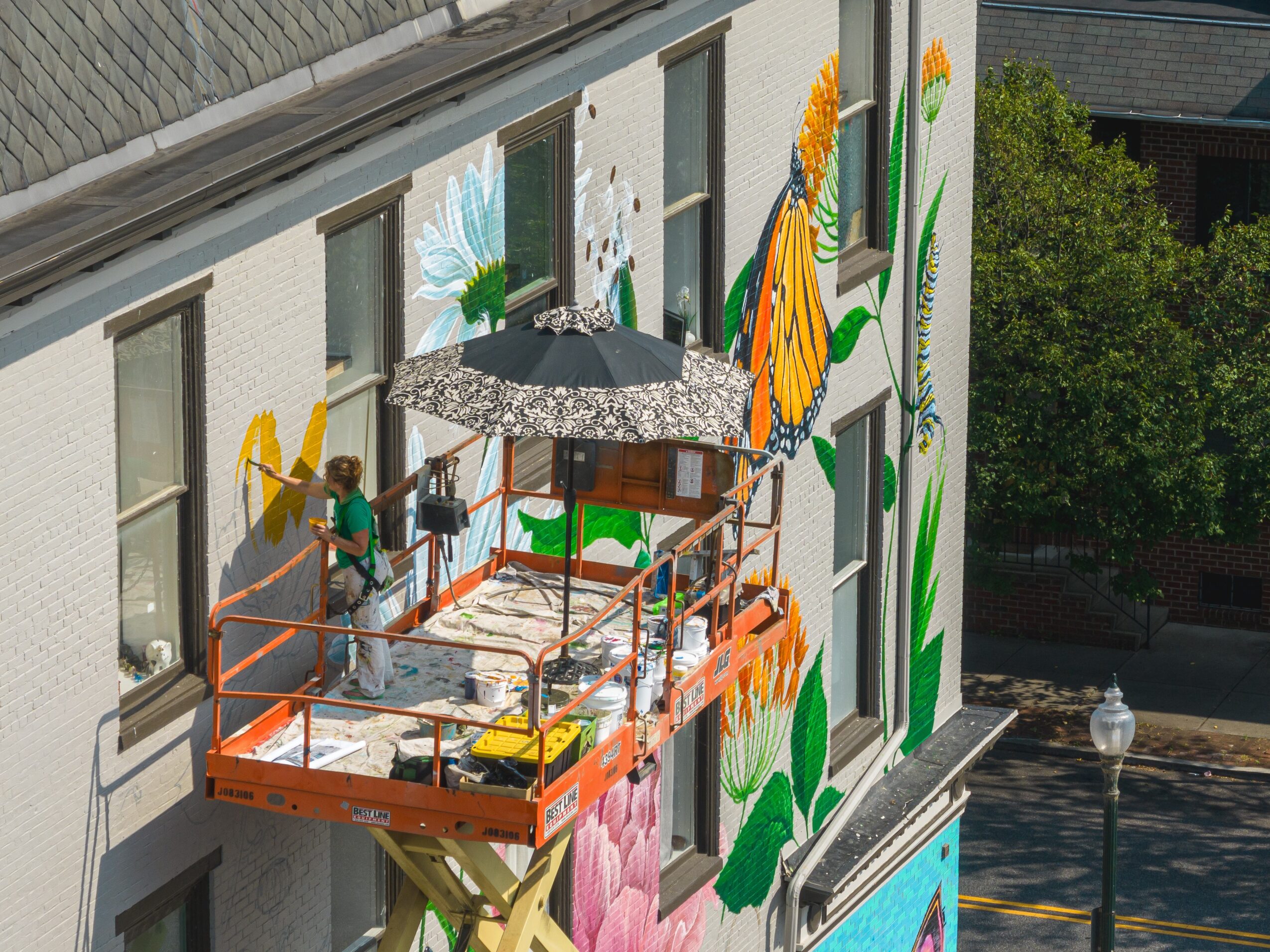 A mural of flowers painted on a building wall with a balcony and a person working on it, seen from below.