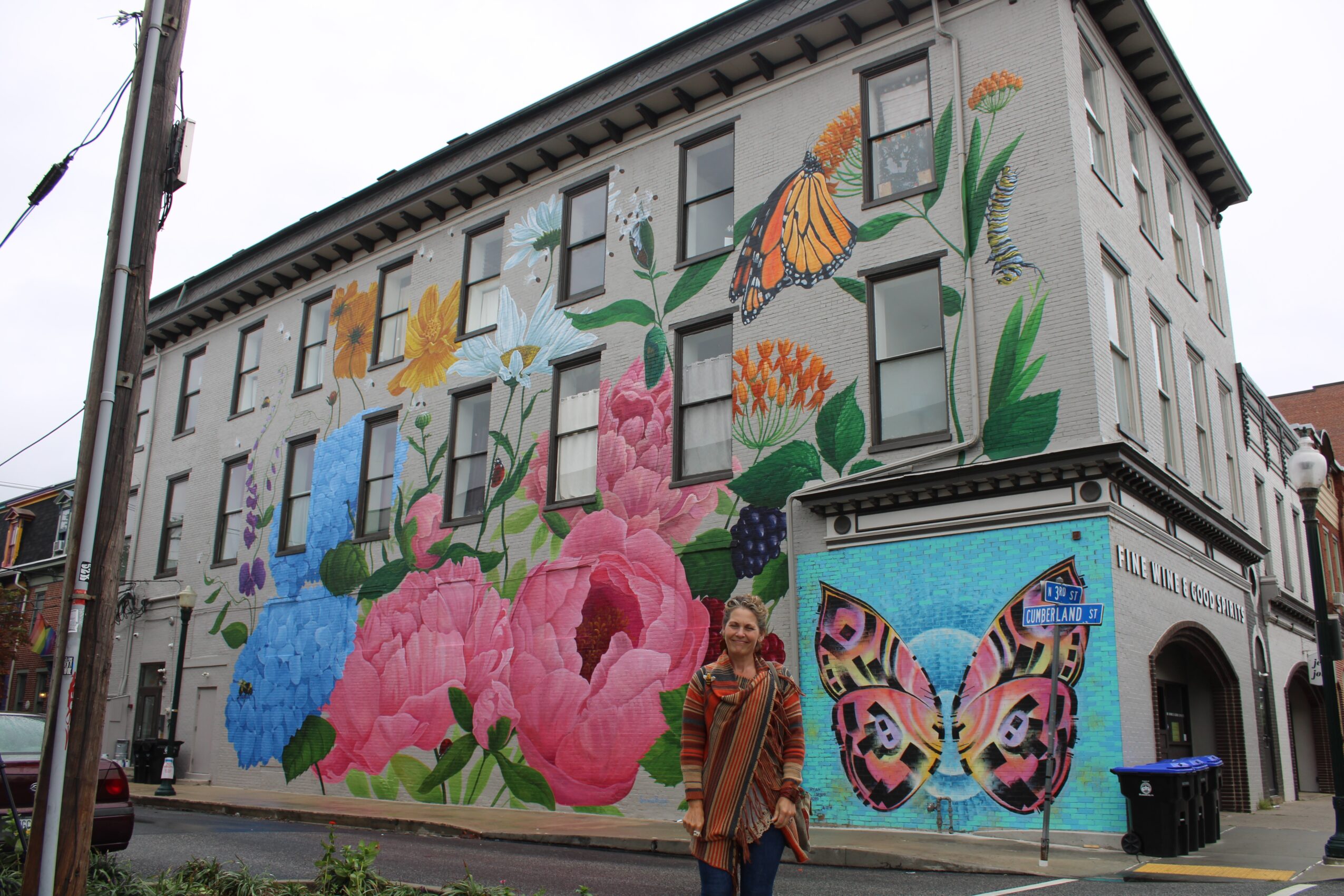 Person standing in front of a building with colorful mural of flowers, butterflies, and plants on the wall.
