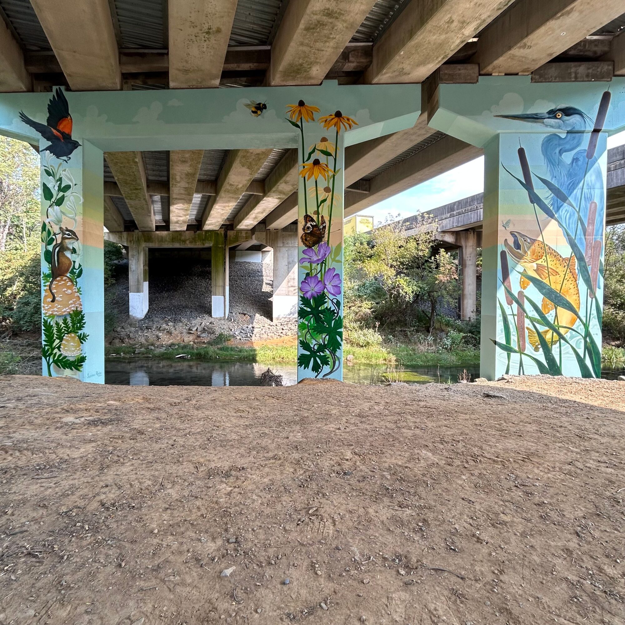 Under a bridge with painted murals of flowers, butterflies, and a dancer, with a river and greenery in the background.