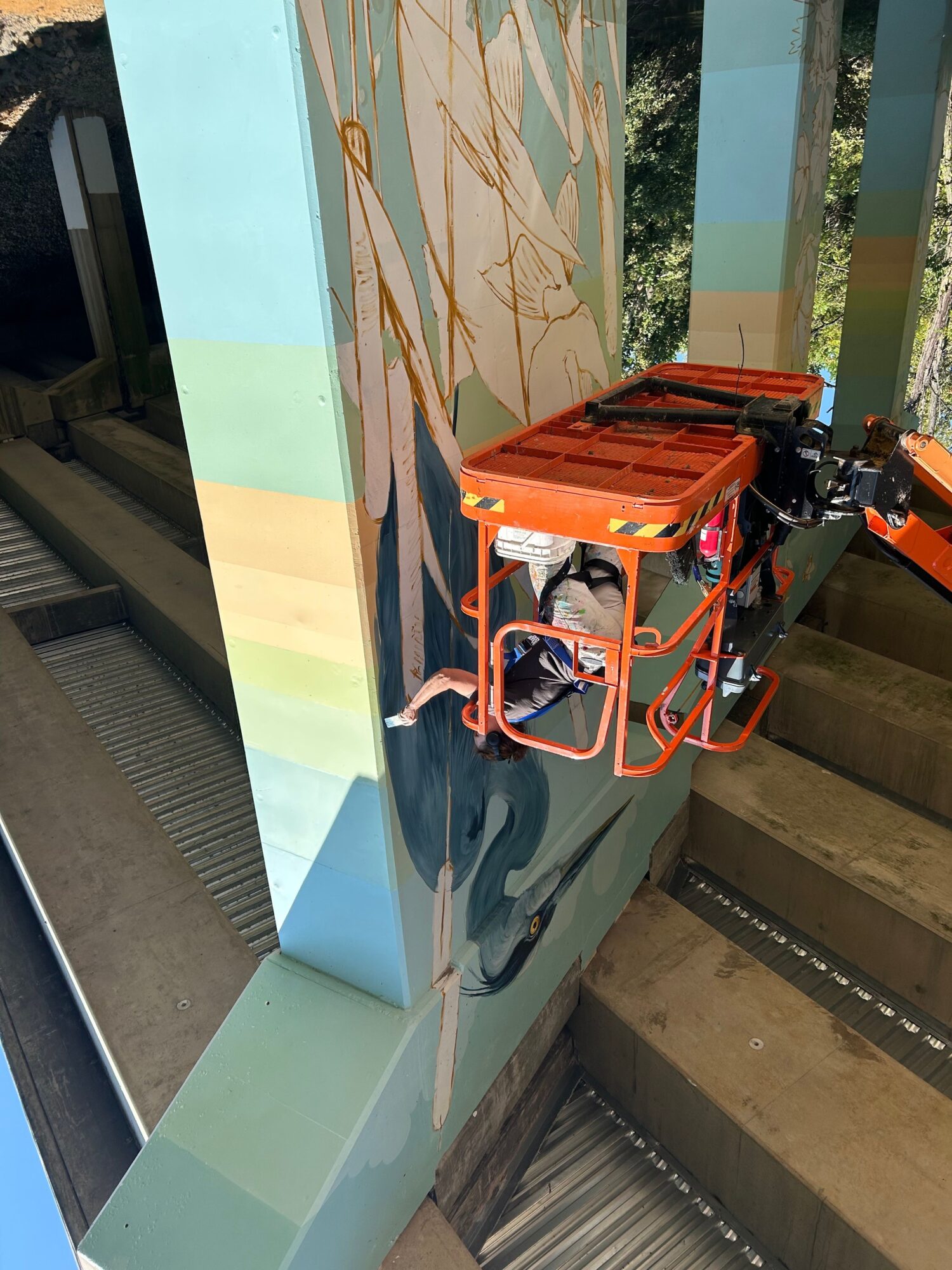 Person on an orange lift working on a large mural with a colorful abstract design, outdoors, with a building in the background.