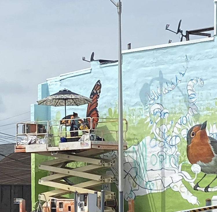 People painting a mural on a building wall with a scissor lift, featuring a bird and colorful designs.