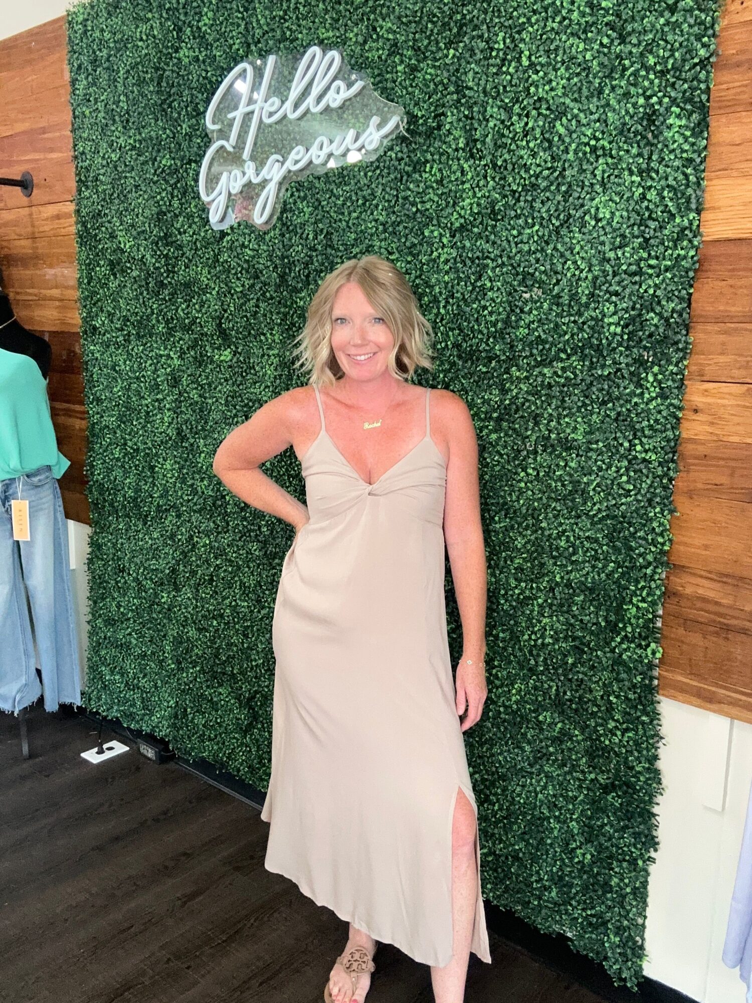 Woman in a light-colored dress standing against a green hedge wall, with a wooden floor and a sign that says 'Hello Gorgeous'.