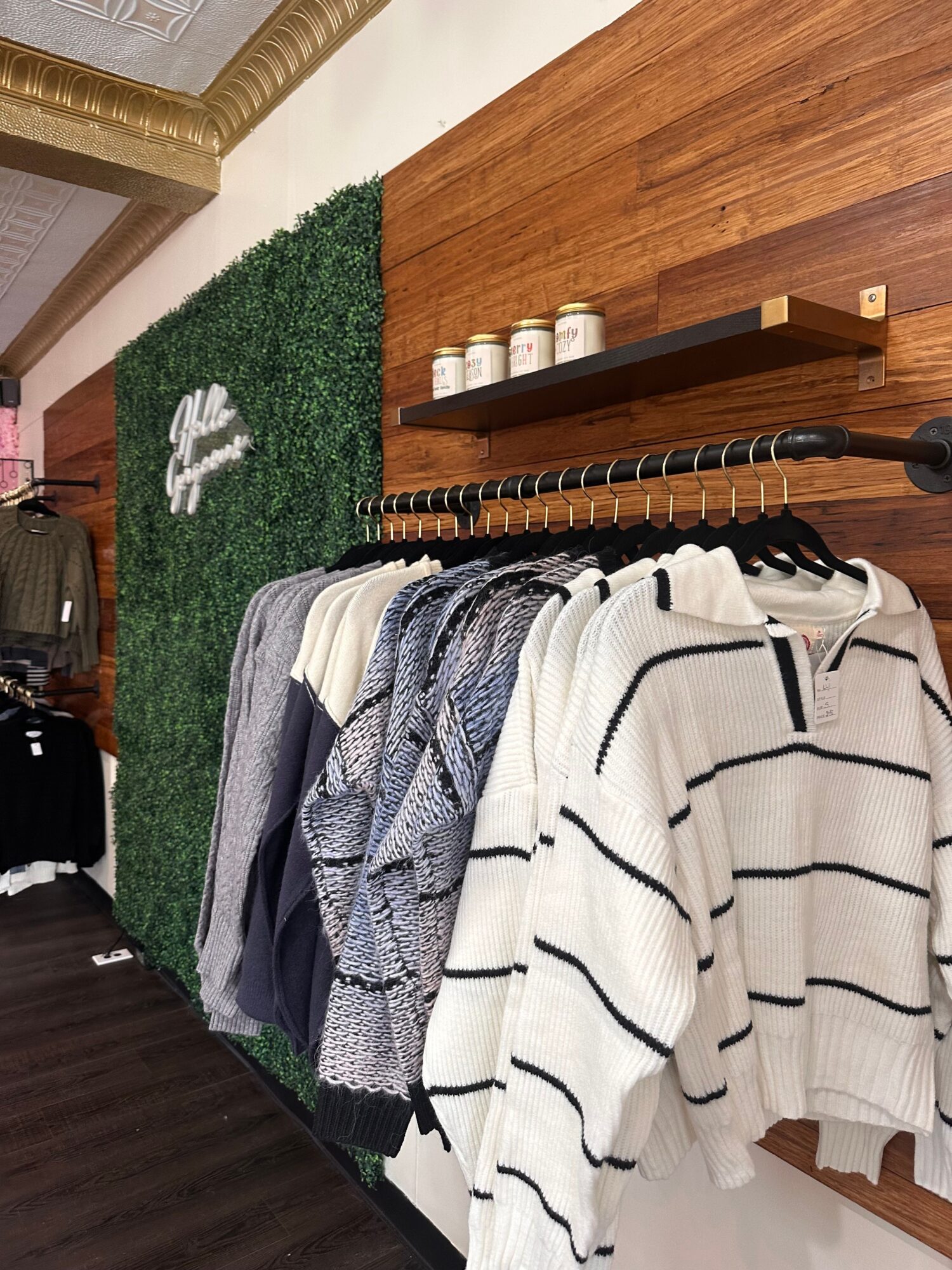 Stacked clothes on a bed with a striped blanket, a green rug with a white logo, and a wooden shelf with bottles.