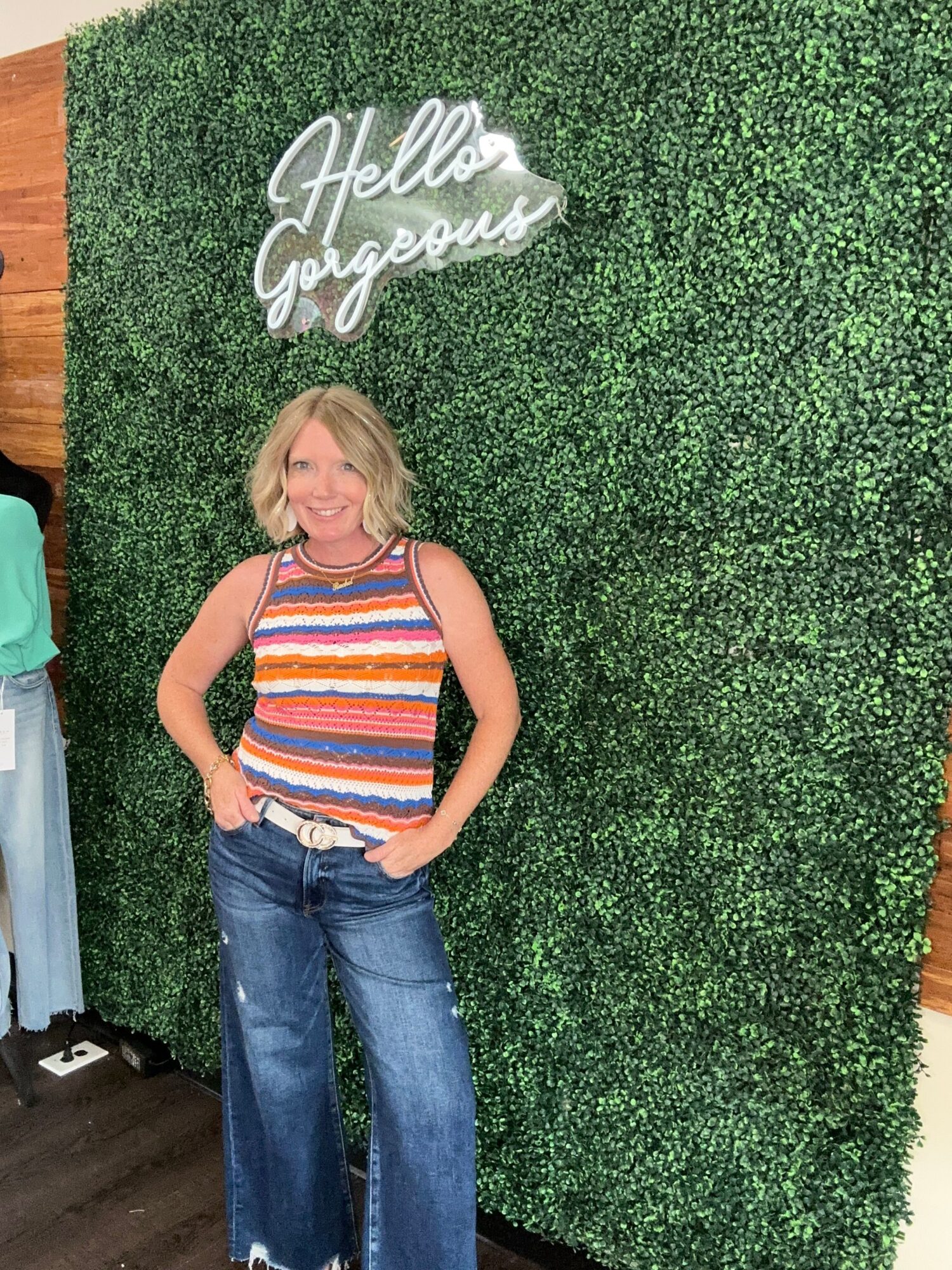 Person with blonde hair wearing a striped shirt and jeans standing against a green hedge wall.
