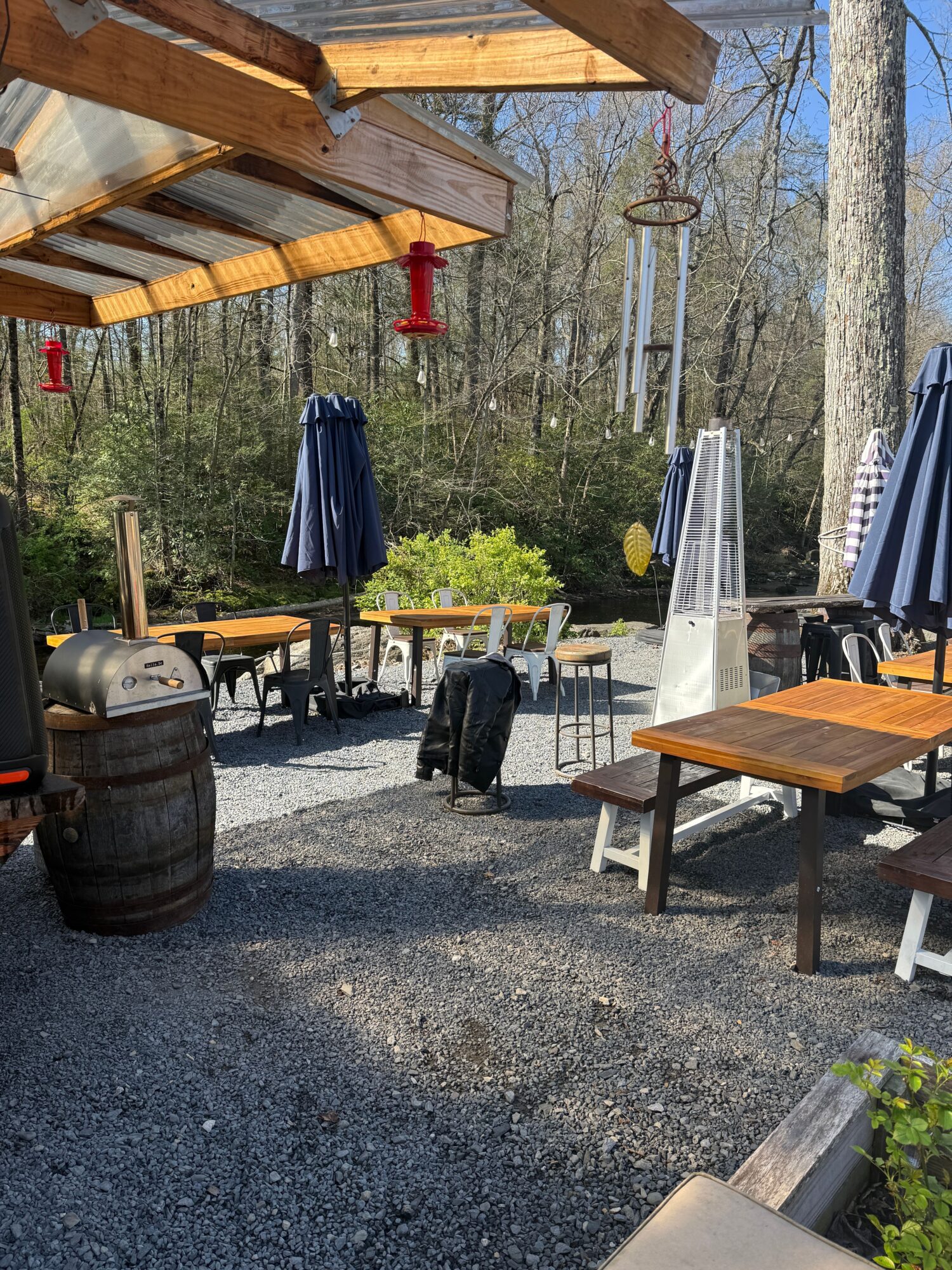 Outdoor patio with tables, chairs, umbrellas, and trees in the background, sunlight, gravel ground, and a barrel in the foreground.