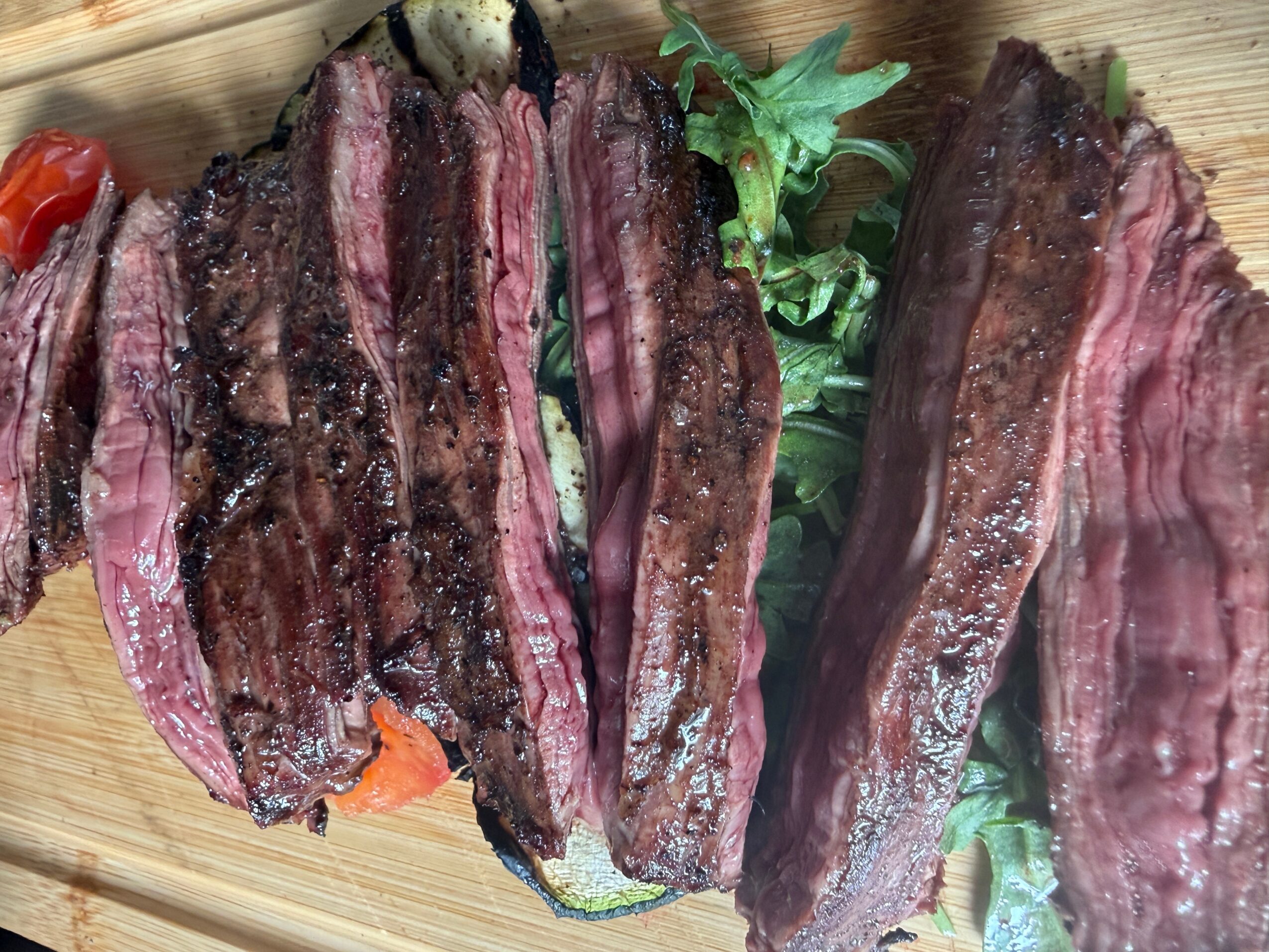 Slices of cooked beef with grill marks on a wooden surface, garnished with greens and vegetables.