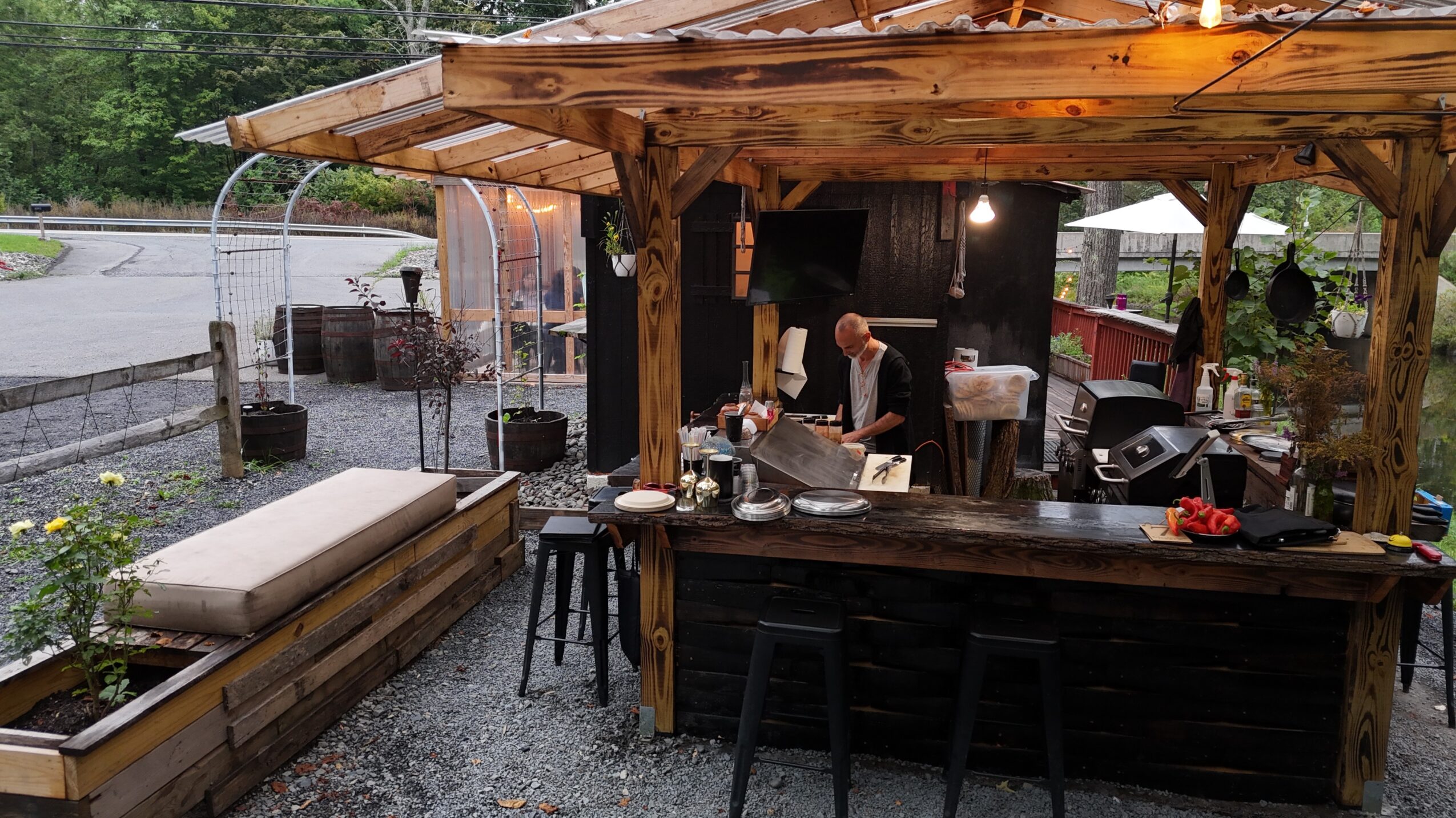 People under a wooden outdoor shelter preparing food, with a bar counter and outdoor seating area, during daytime.