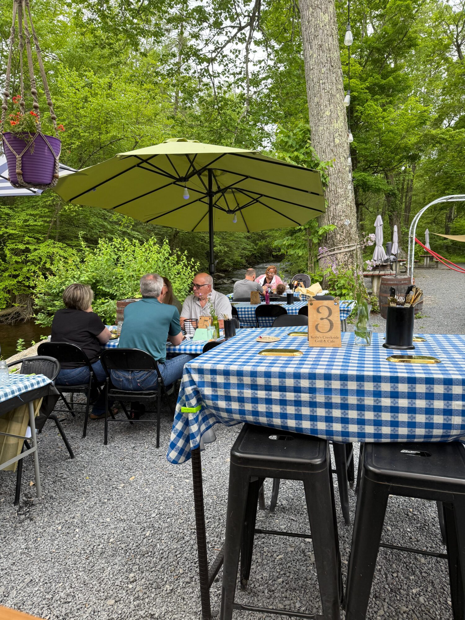 People sitting at a table outdoors under a green umbrella, with trees and greenery in the background.