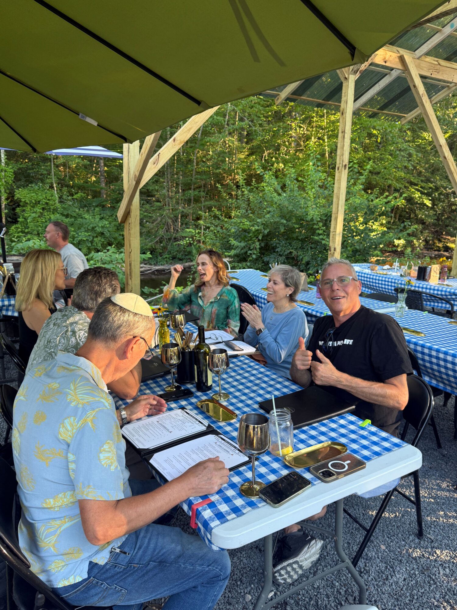 People sitting at a table outdoors with a blue and white checkered tablecloth, drinks, and a camera, surrounded by greenery.