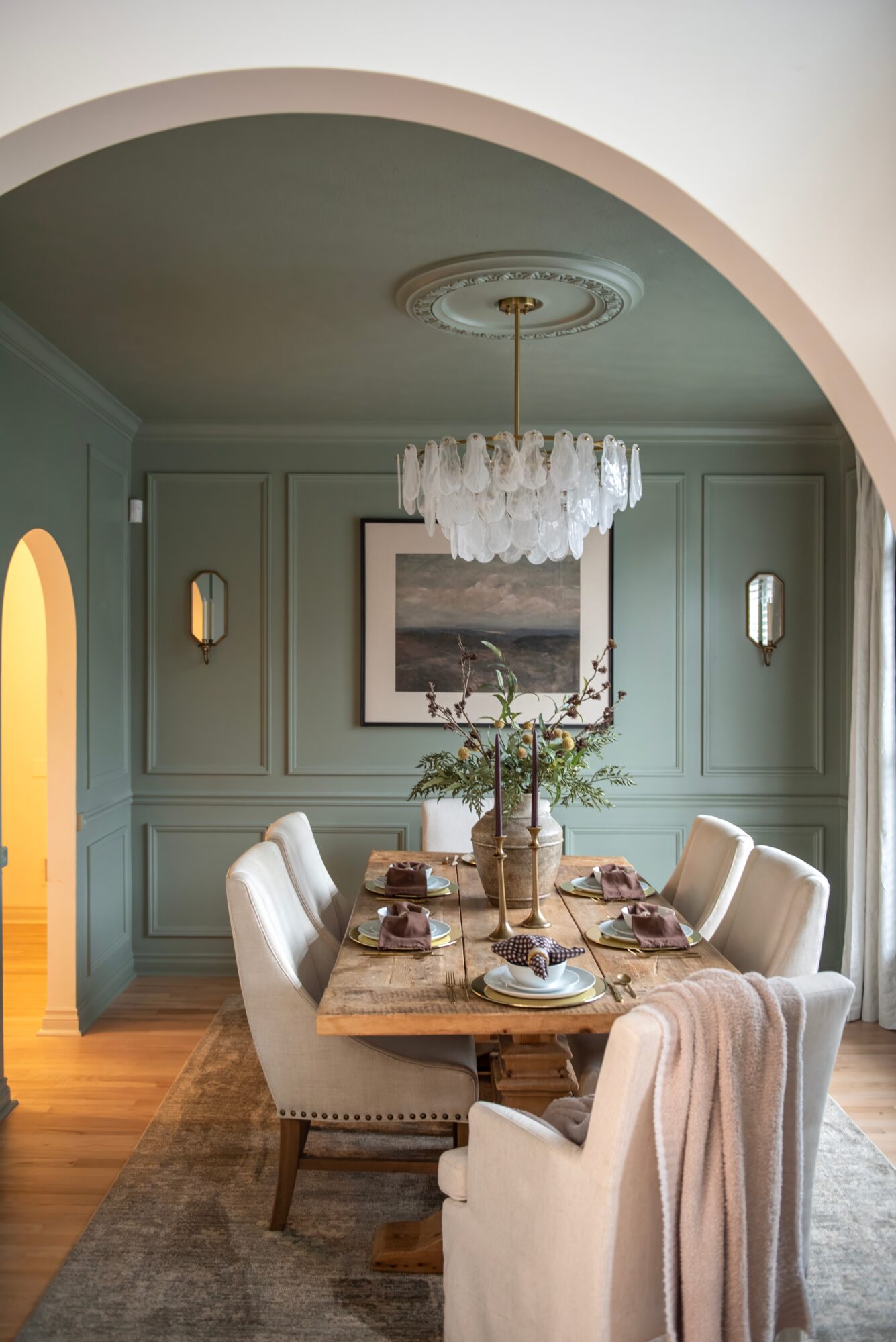 Dining room with a wooden table, six white chairs, a chandelier, and a framed picture on a green wall.