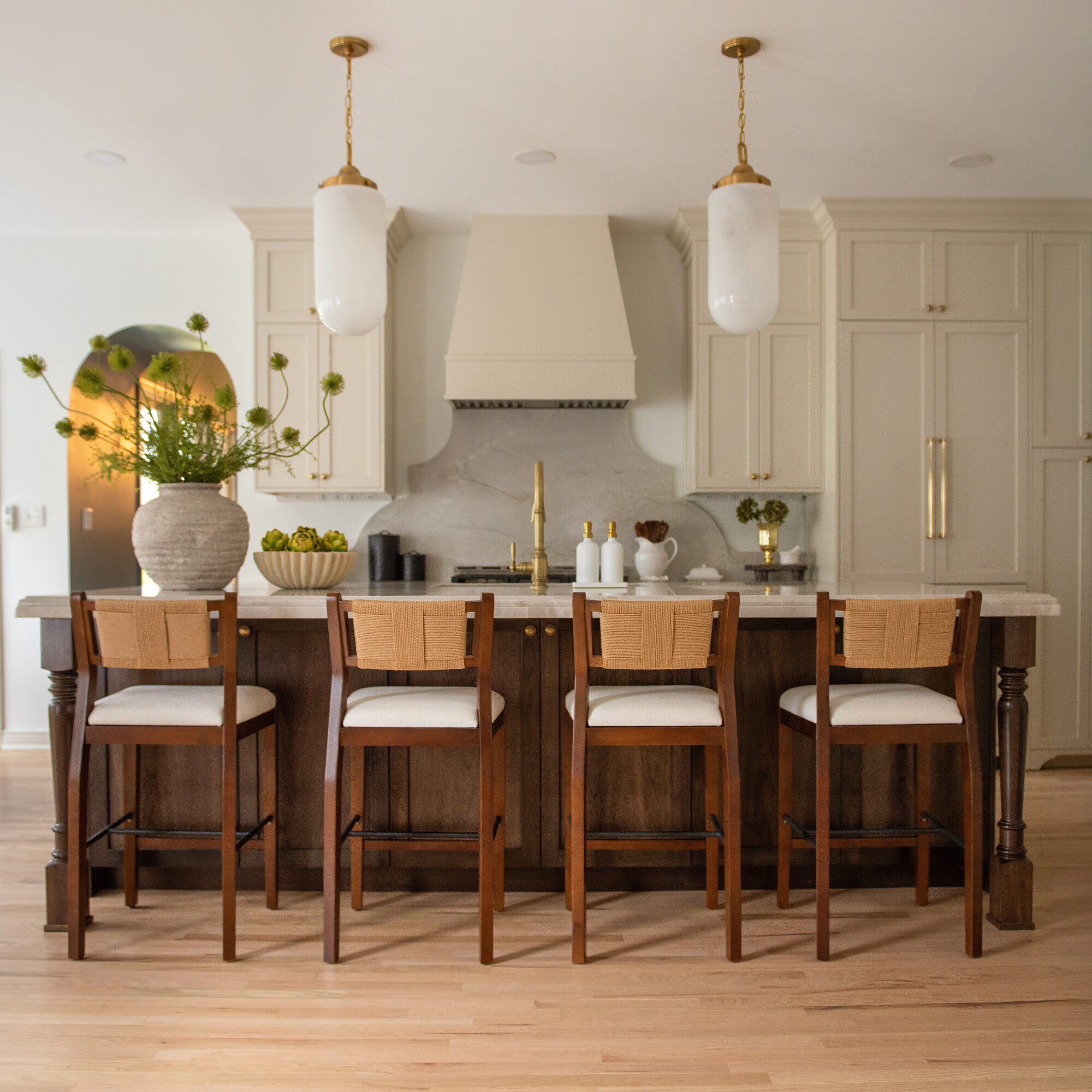 Kitchen with four wooden bar stools at a dark wood island, white cabinets, and two pendant lights, with a vase and fruit on the counter.