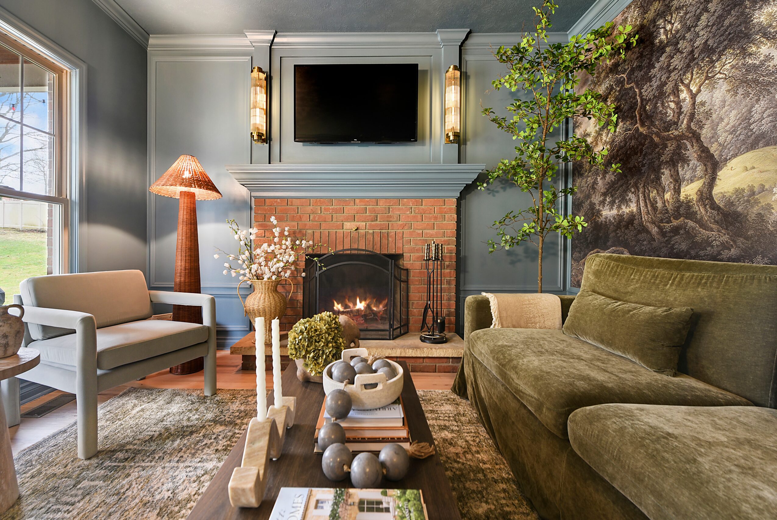 Living room with fireplace, TV above, green sofa, armchair, and decorative plants, with a large landscape mural on the wall.