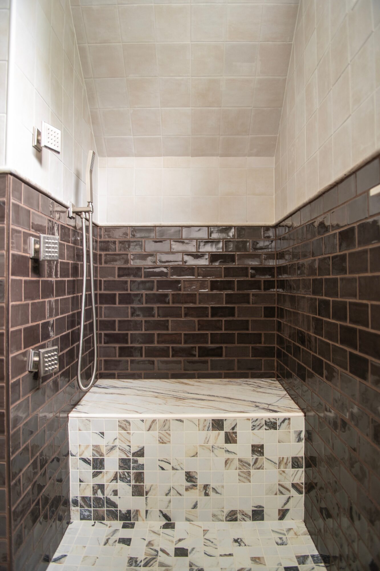 Small shower area with beige and dark brown tiled walls, marble bench, and a handheld showerhead.
