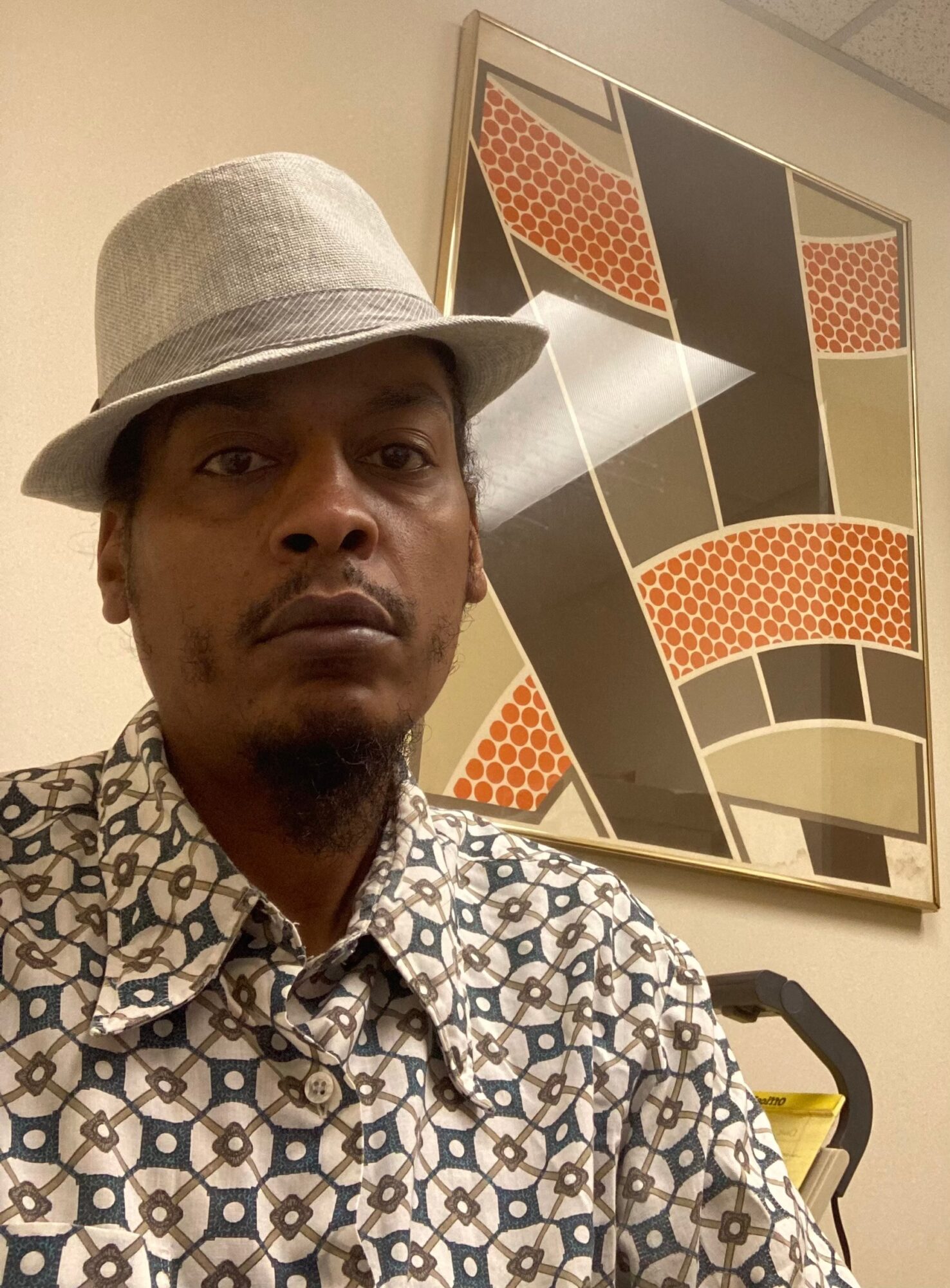 Person wearing a light-colored hat and patterned shirt, standing in front of geometric artwork on a wall.