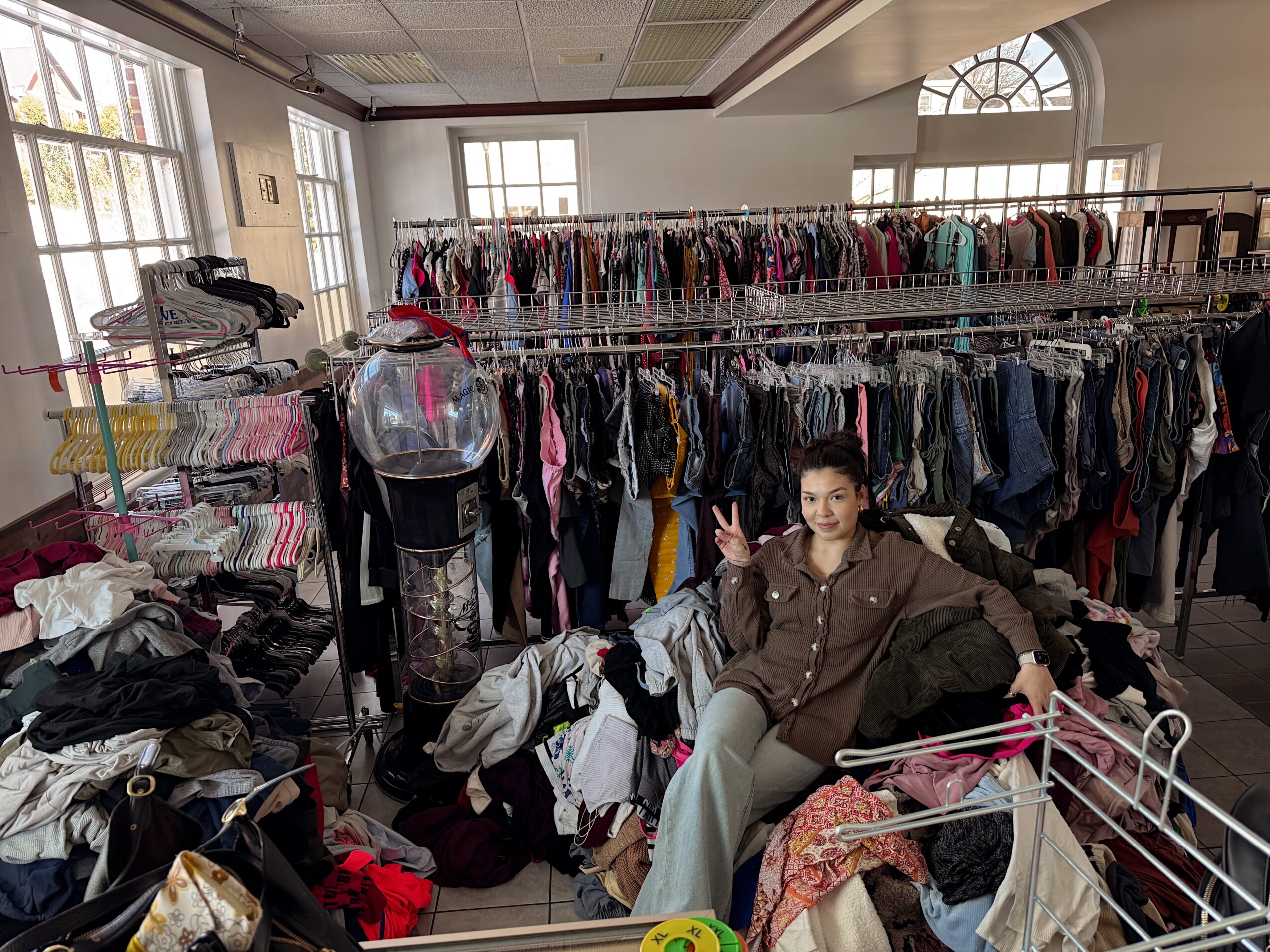 Person sitting among clothing racks in a store, making a peace sign with one hand, surrounded by clothes and shopping carts.