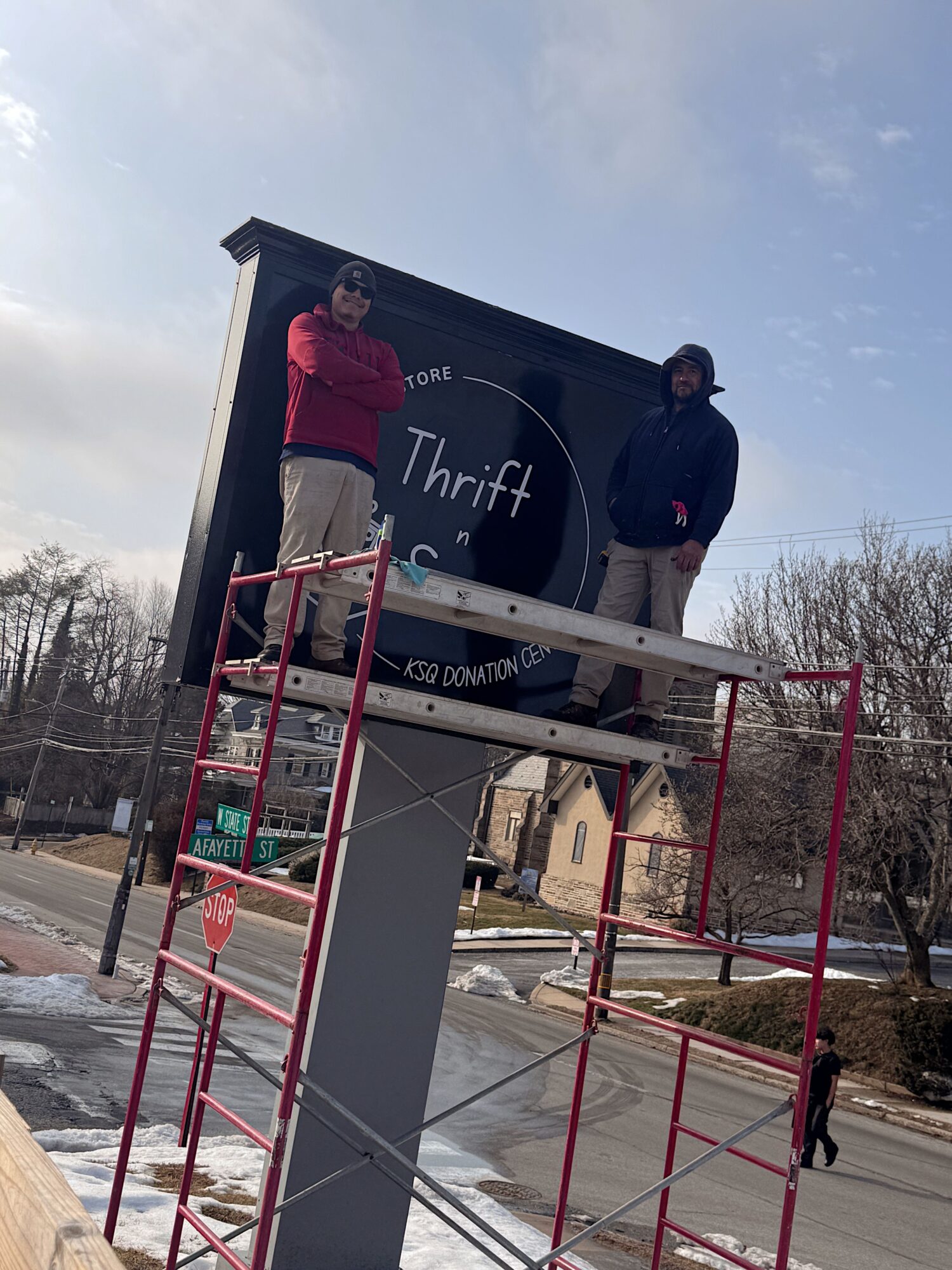 Person standing on a ladder in front of a Thrift sign, with a cloudy sky and trees in the background.