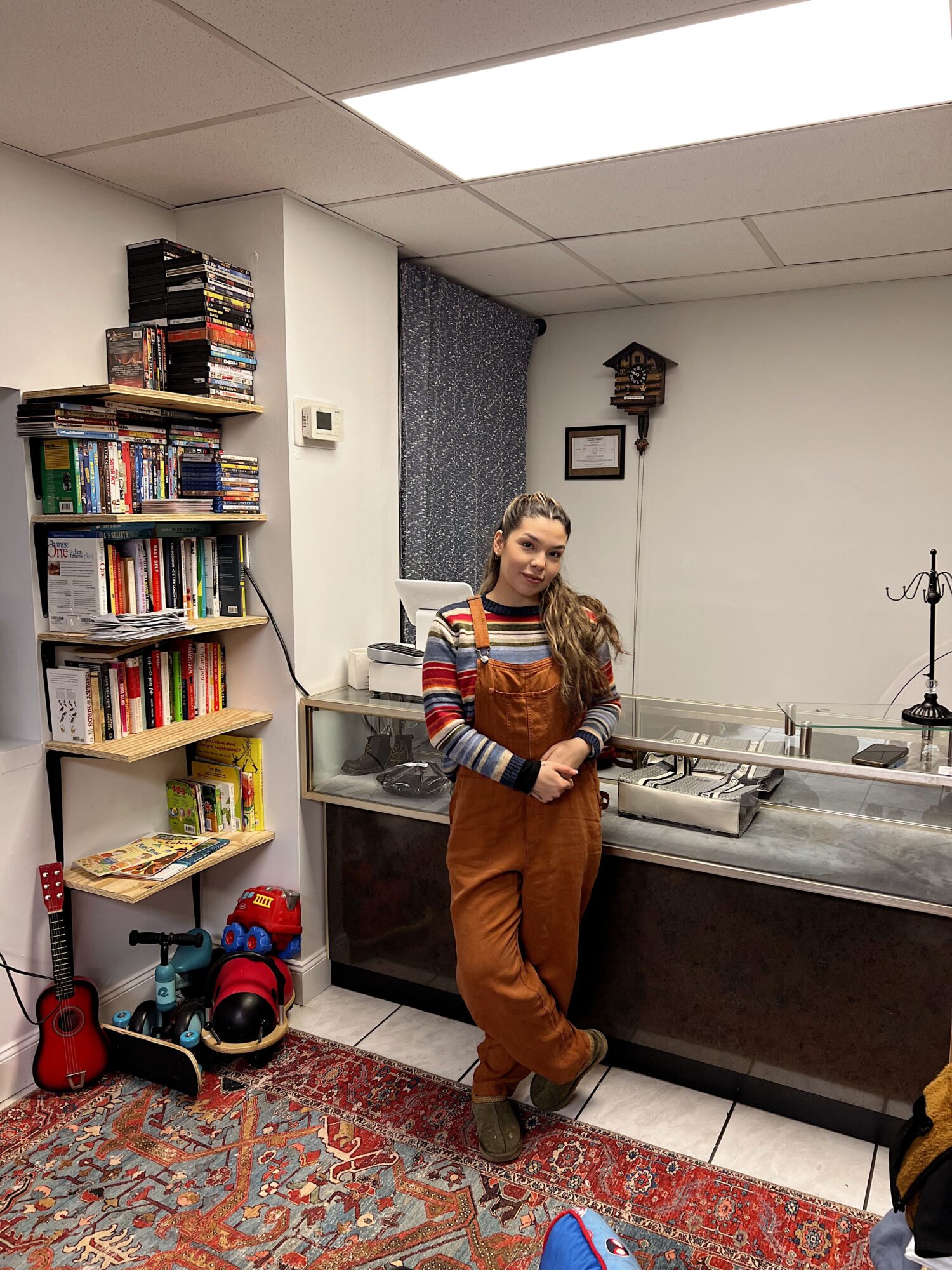 Person standing in a kitchen, holding a mug, with bookshelves and a colorful rug nearby.