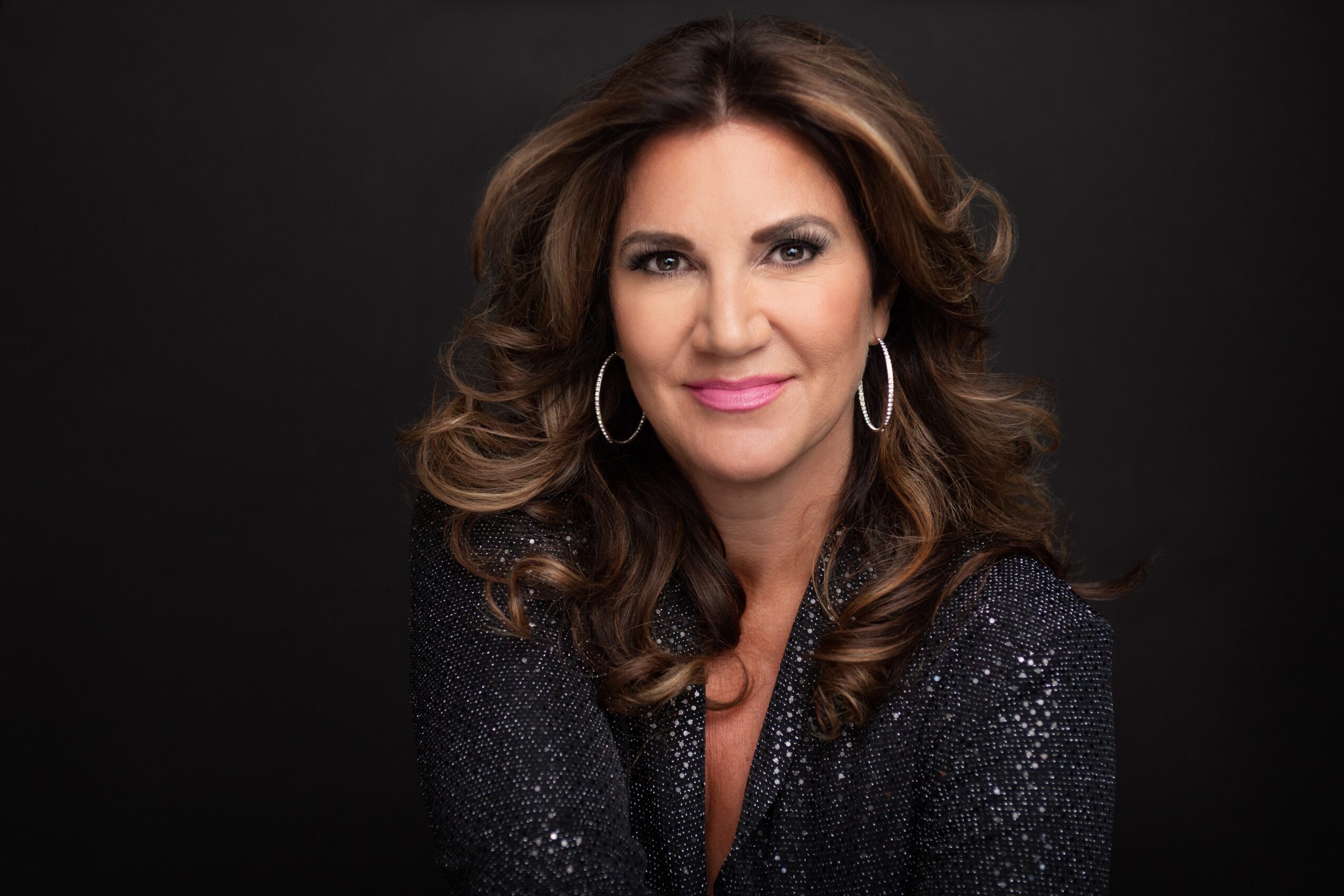 Woman with wavy brown hair, wearing hoop earrings and a dark, sparkly top, smiling against a black background.