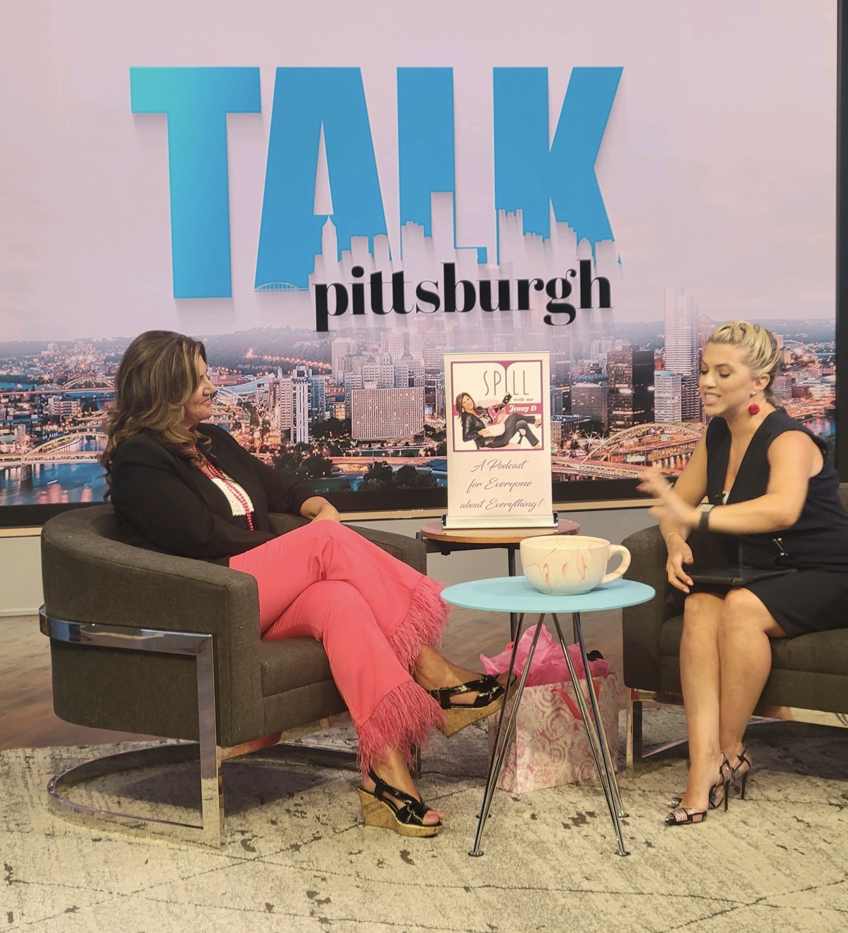 Two women sit on sofas in a talk show set with a cityscape background, a small table with a bowl, and a framed picture.