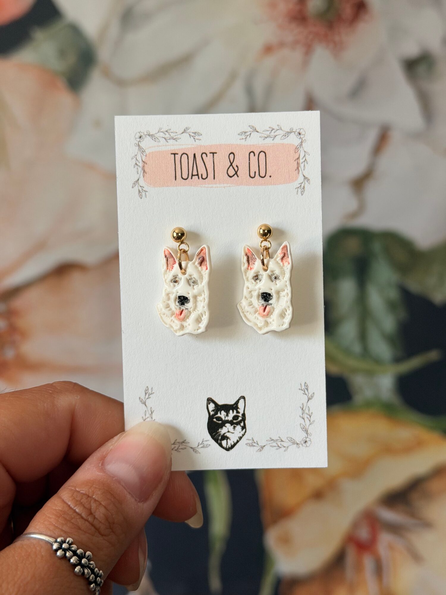 Hand holding a card with two small animal-shaped earrings and a black cat illustration, background of floral fabric.