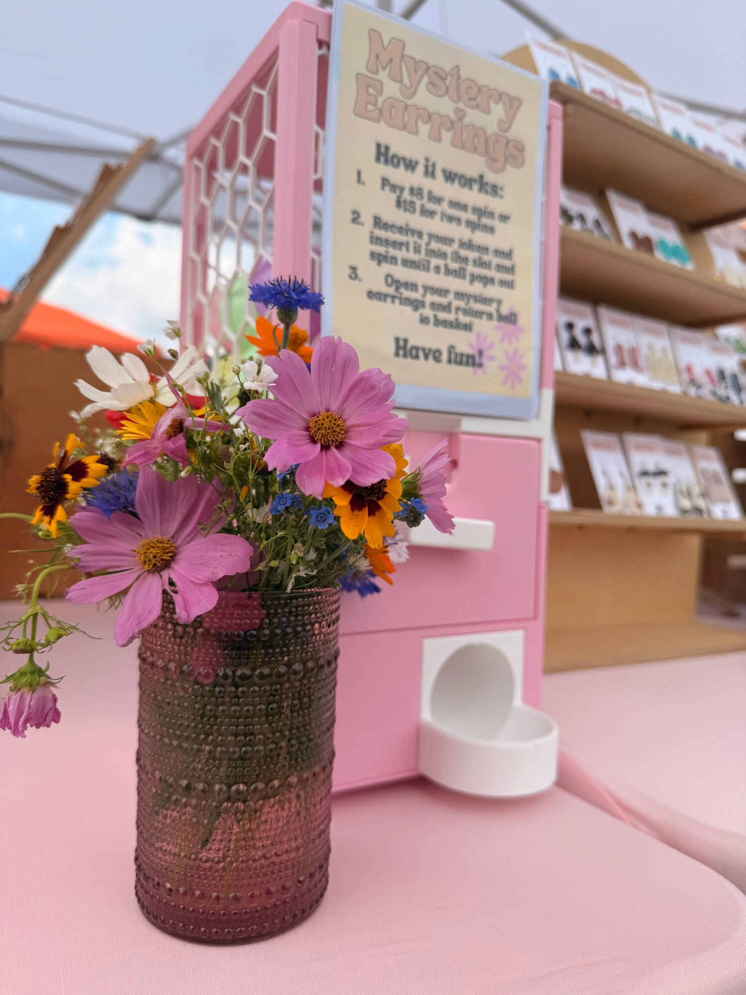 Pink flower arrangement in a dark vase on a pink surface with a white heart-shaped container and a sign in the background.
