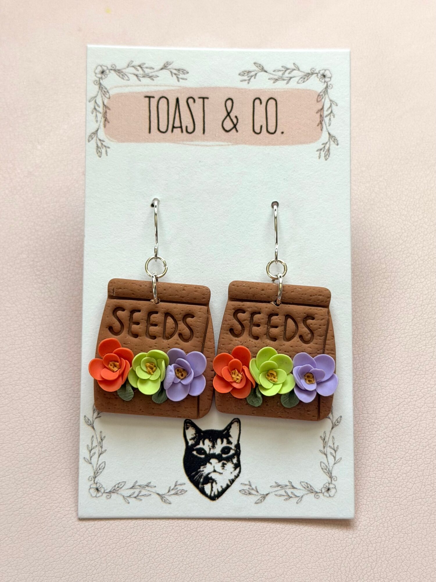 Earrings with seed packet and flower designs hang on a card with 'Toast & Co.' and a cat illustration.