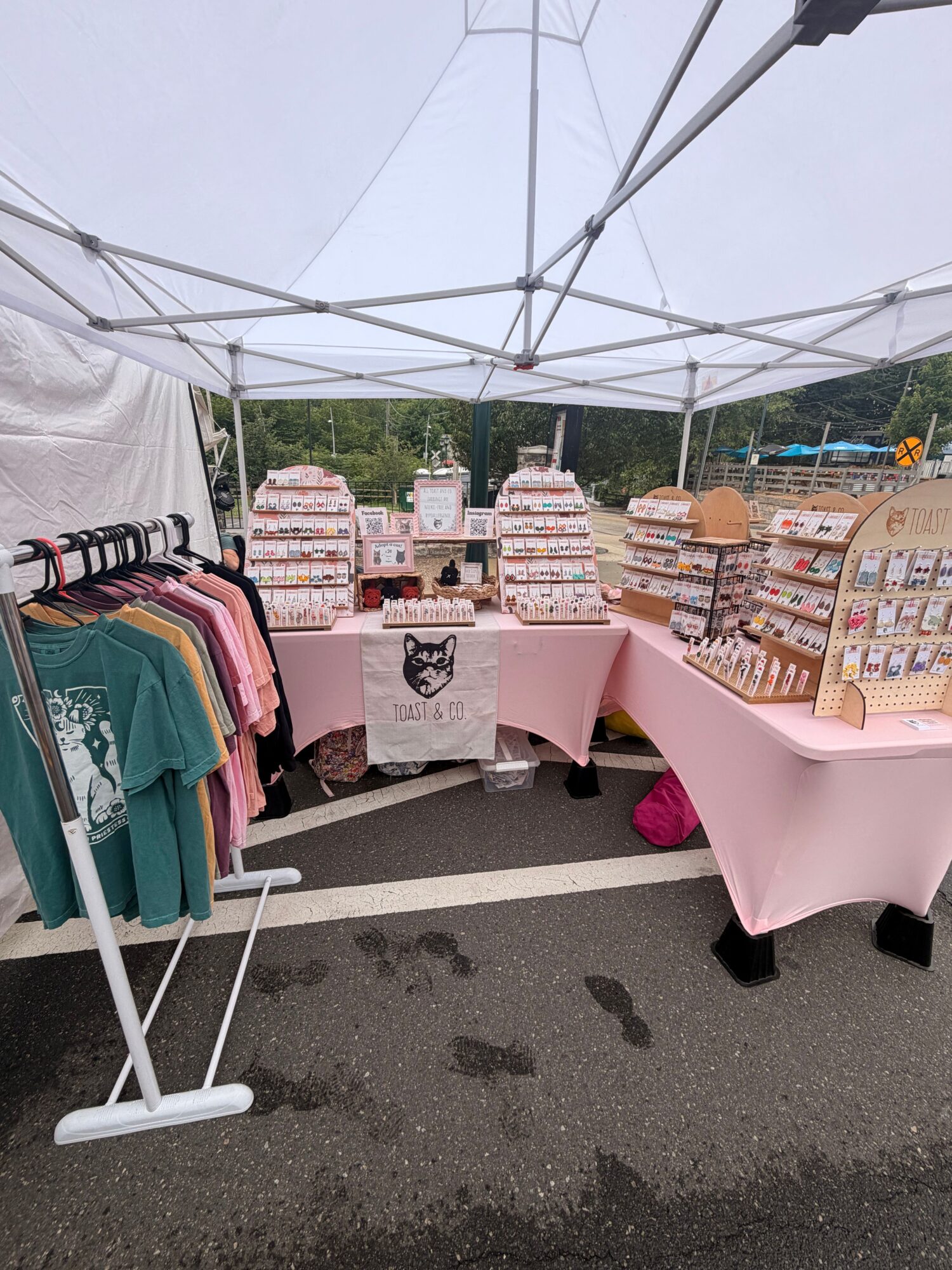 Outdoor market stall with pink tablecloth, clothing, and boxes under a canopy, on a paved street.
