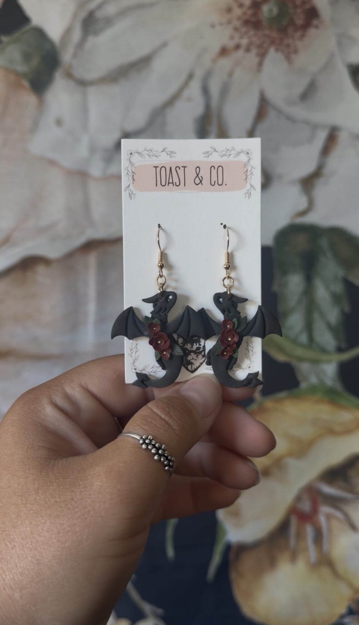 Hand holding earrings with bat and flower designs on a white card, background of flowers and leaves.