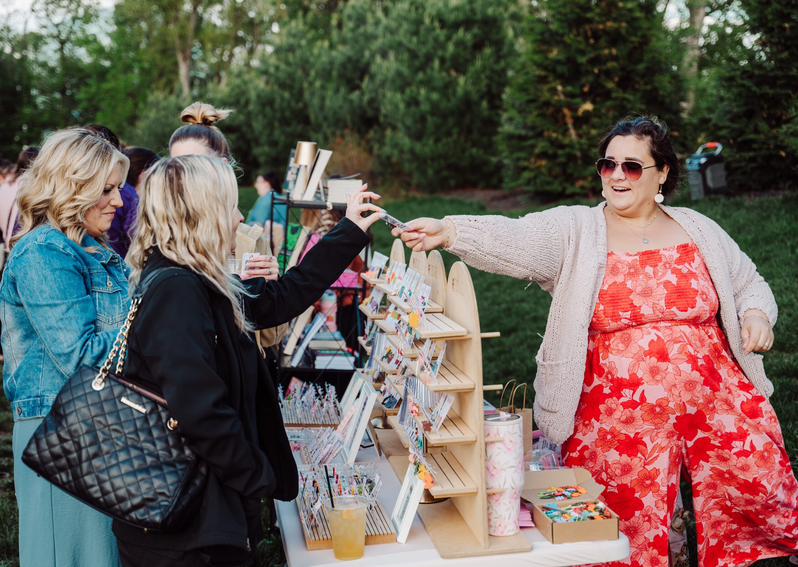 Woman in red floral outfit and sunglasses hands a drink to a woman at an outdoor market stall, with other people nearby.