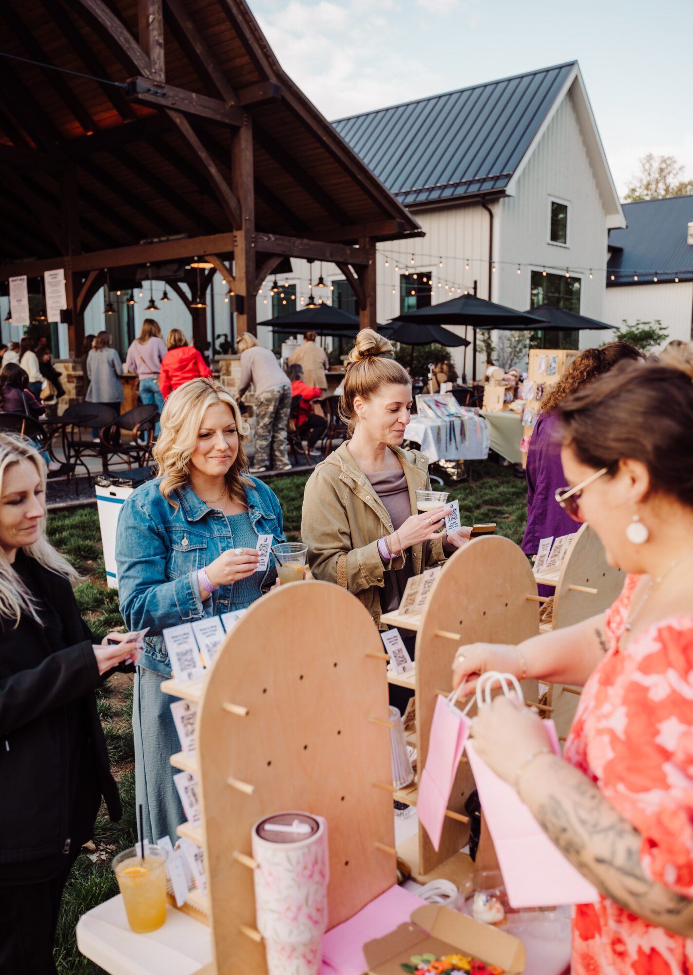 People gather outdoors at a social event with tables, chairs, and a building in the background, some holding drinks.