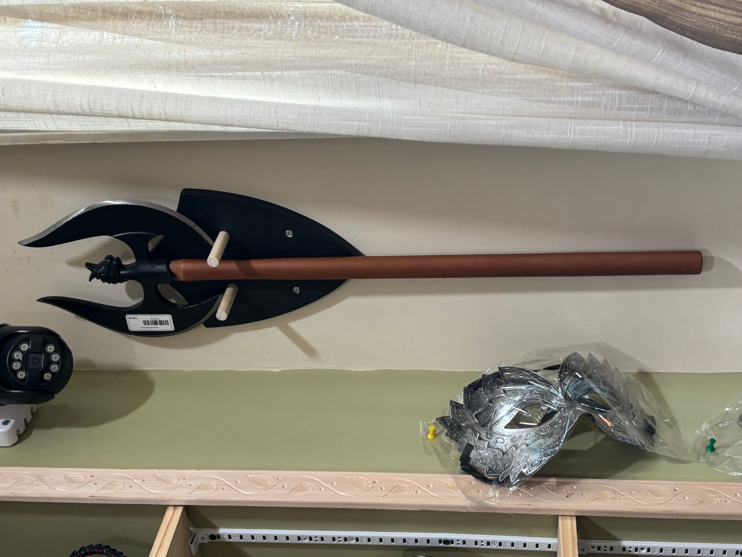 A black spear with a wooden shaft and a metal spearhead is mounted on a wall, with a plastic bag and a remote control on a shelf below.