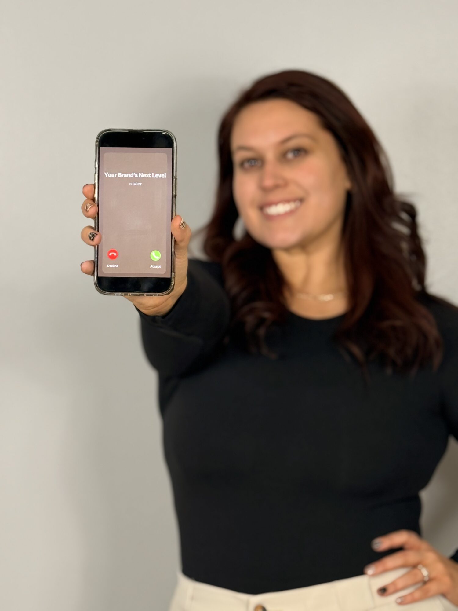 Woman smiling and holding a phone showing an incoming call, standing against a plain background.