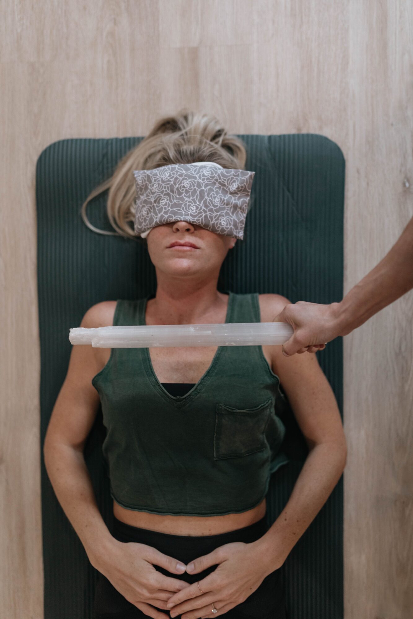 Person lying on a yoga mat with a cloth over their eyes, holding a cylindrical object, in a wellness setting.