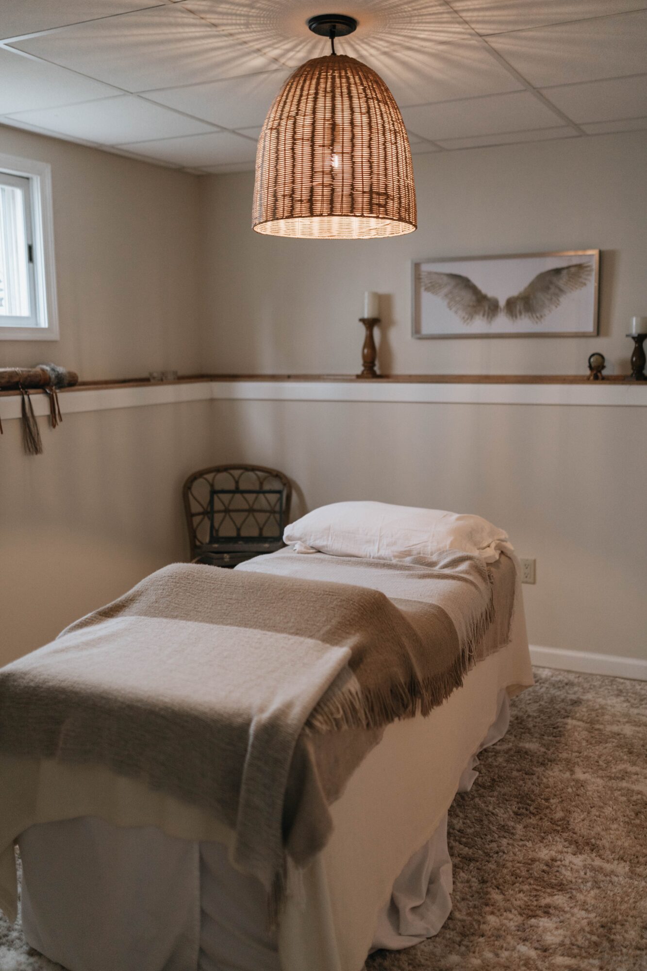 A cozy bedroom with a bed, wicker pendant light, framed butterfly artwork, and decorative candles on a shelf.