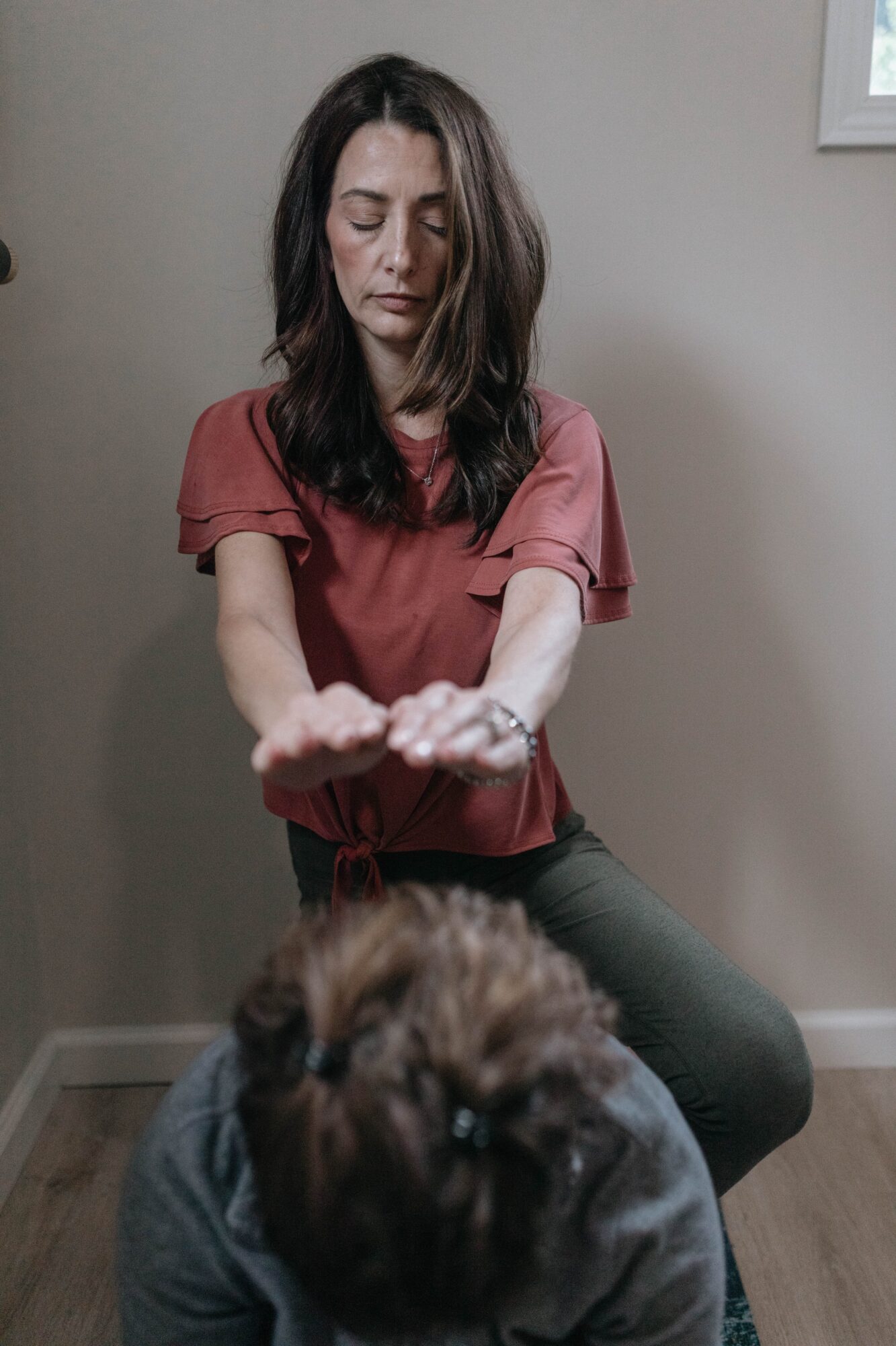 Woman with long dark hair in a pink top extends her arms forward, with a dog in front of her, indoors.