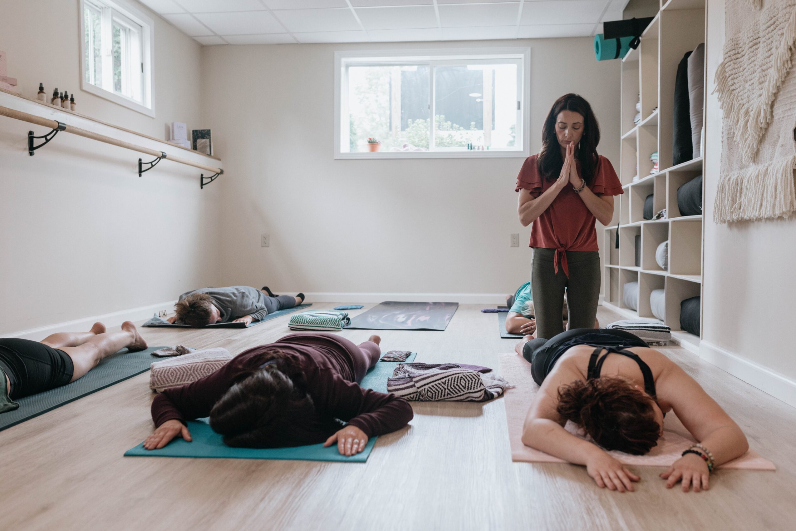 Four people lying on yoga mats in a bright room, with a woman standing and praying, and shelves on the wall.