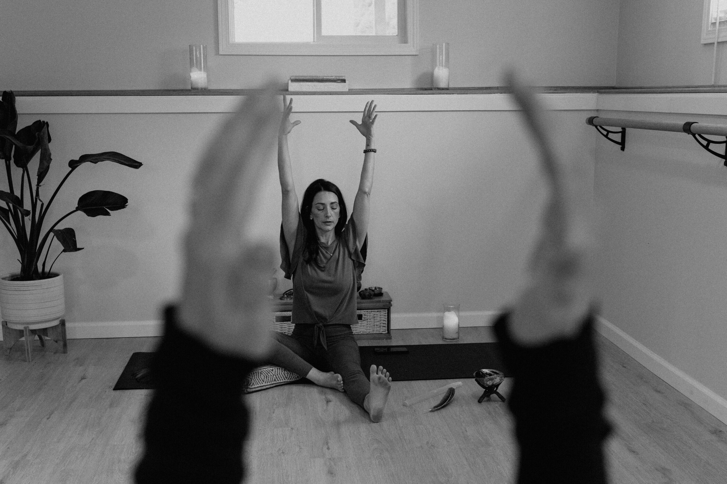 A woman practicing yoga in a room with two people in the foreground raising their arms, and a plant on the left.
