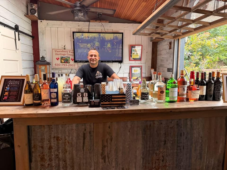 Man standing behind a bar counter with various bottles and glasses, smiling, in a rustic setting with a TV and window.