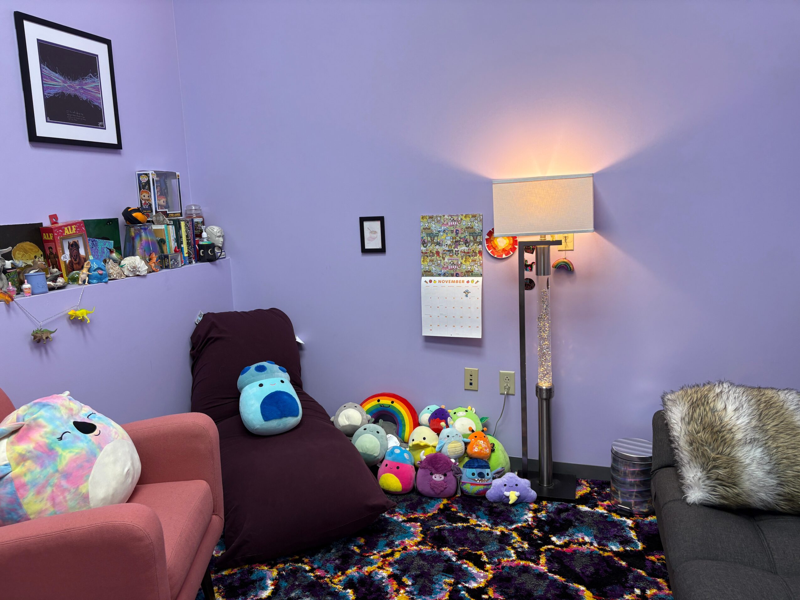 Living room with purple wall, lamp, and colorful plush toys on the floor, with a pink armchair and a gray sofa.
