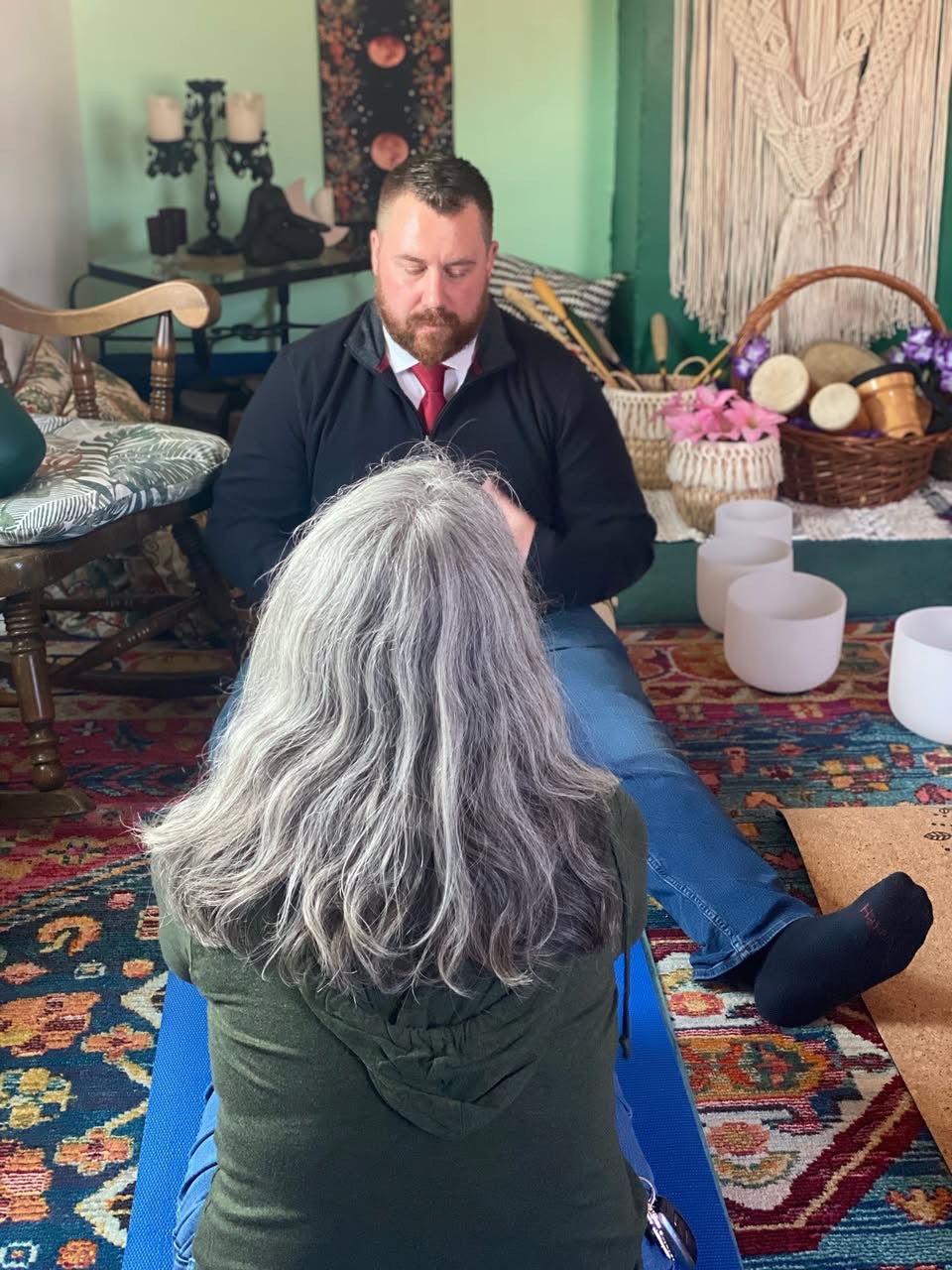 A man with a beard and a woman with long gray hair sit facing each other in a room decorated with baskets, candles, and a colorful rug.