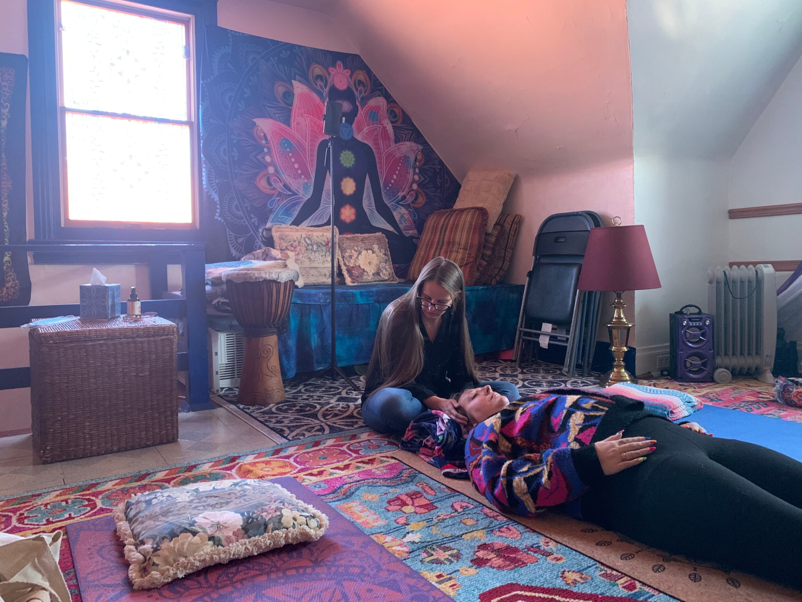 Person lying on colorful blanket on floor, with another person sitting nearby, in a room with a window, tapestry, and furniture.
